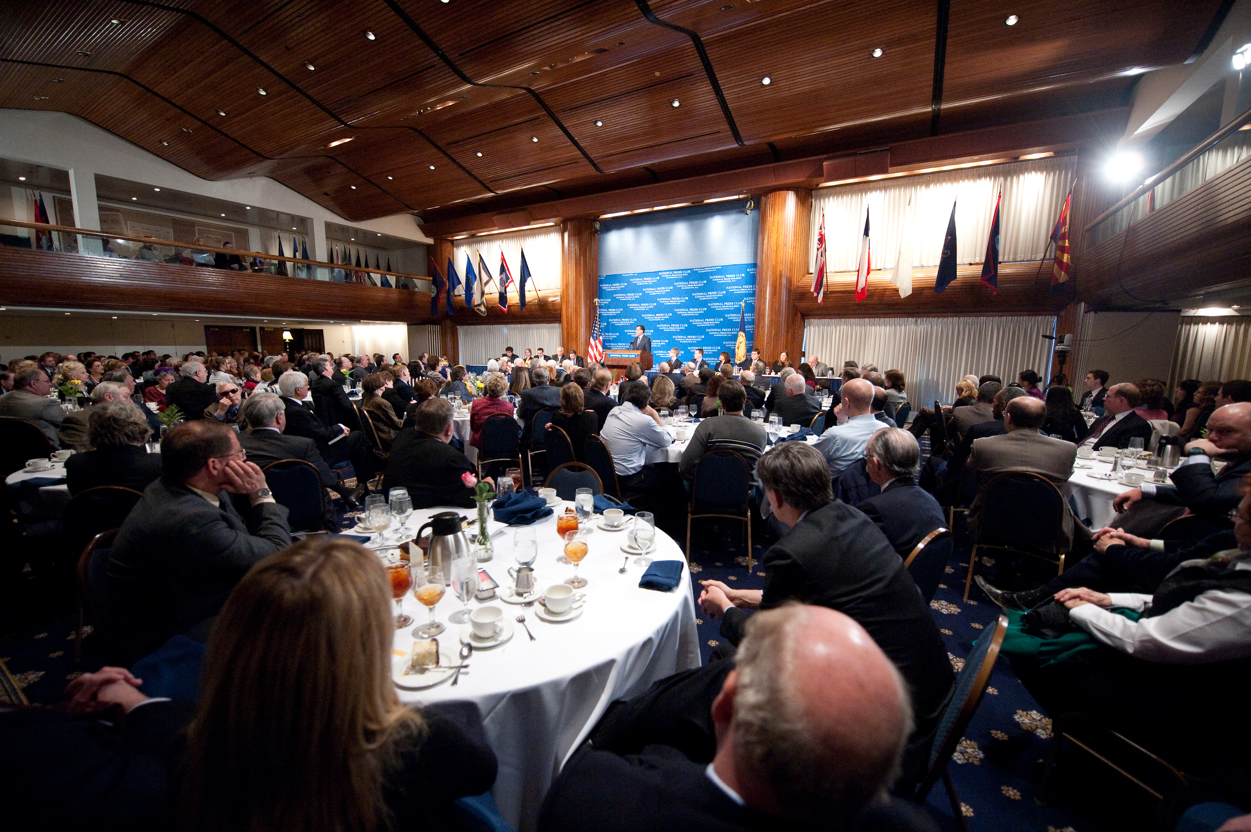 A large audience attends a formal event in a spacious hall with a stage, blue backdrop, and several national flags displayed. People are seated at round tables set with meals.