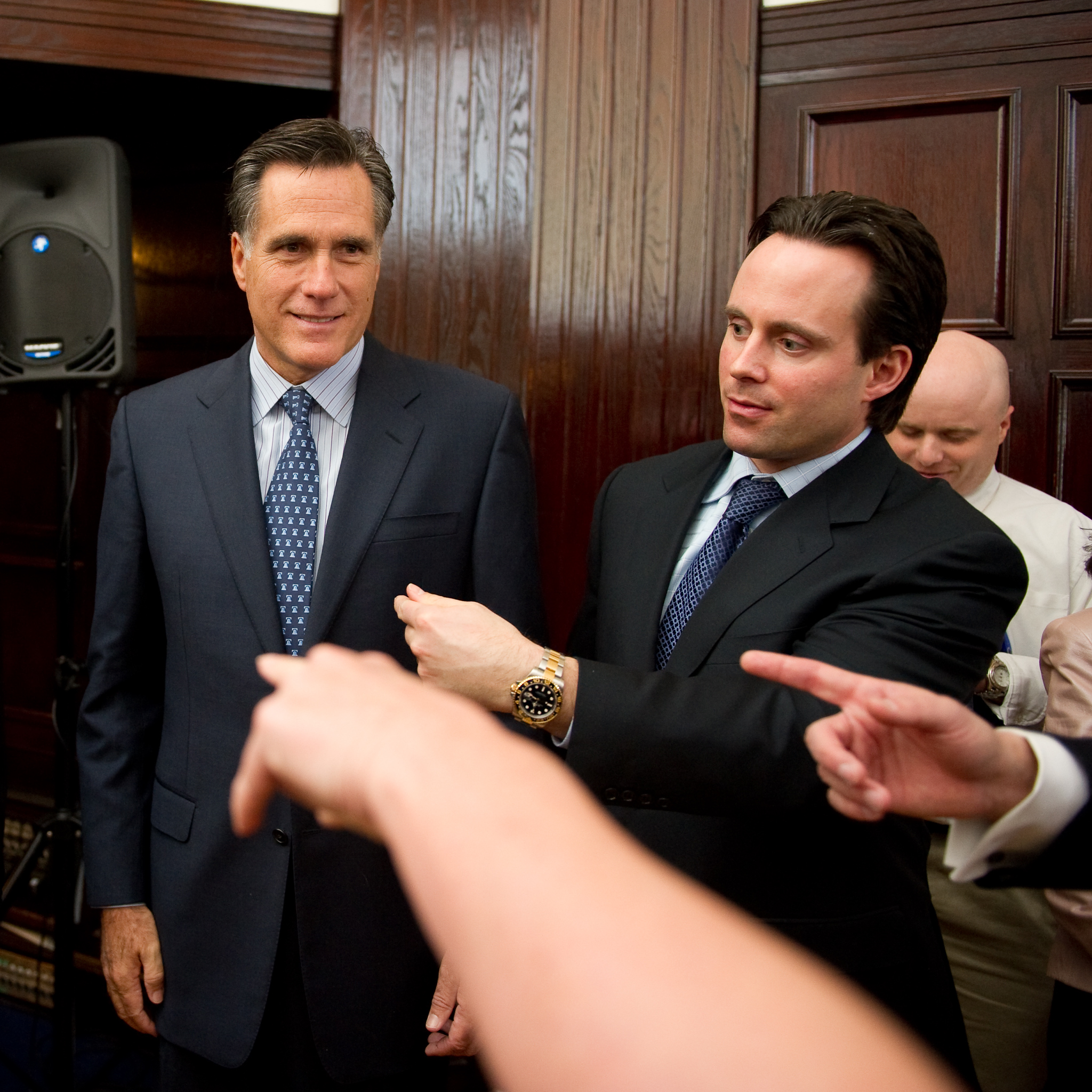 Two men in suits stand in a room with wood-paneled walls, while several hands in the foreground gesture and point toward them.