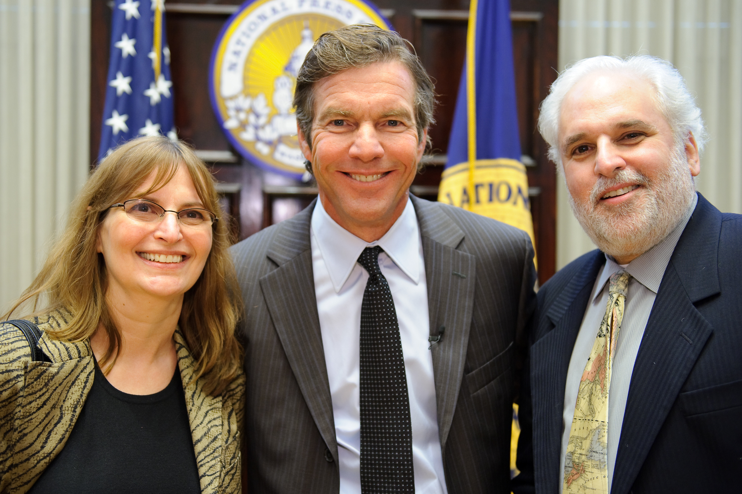 Three adults in business attire smile for a photo together indoors, with flags and an official seal visible in the background.