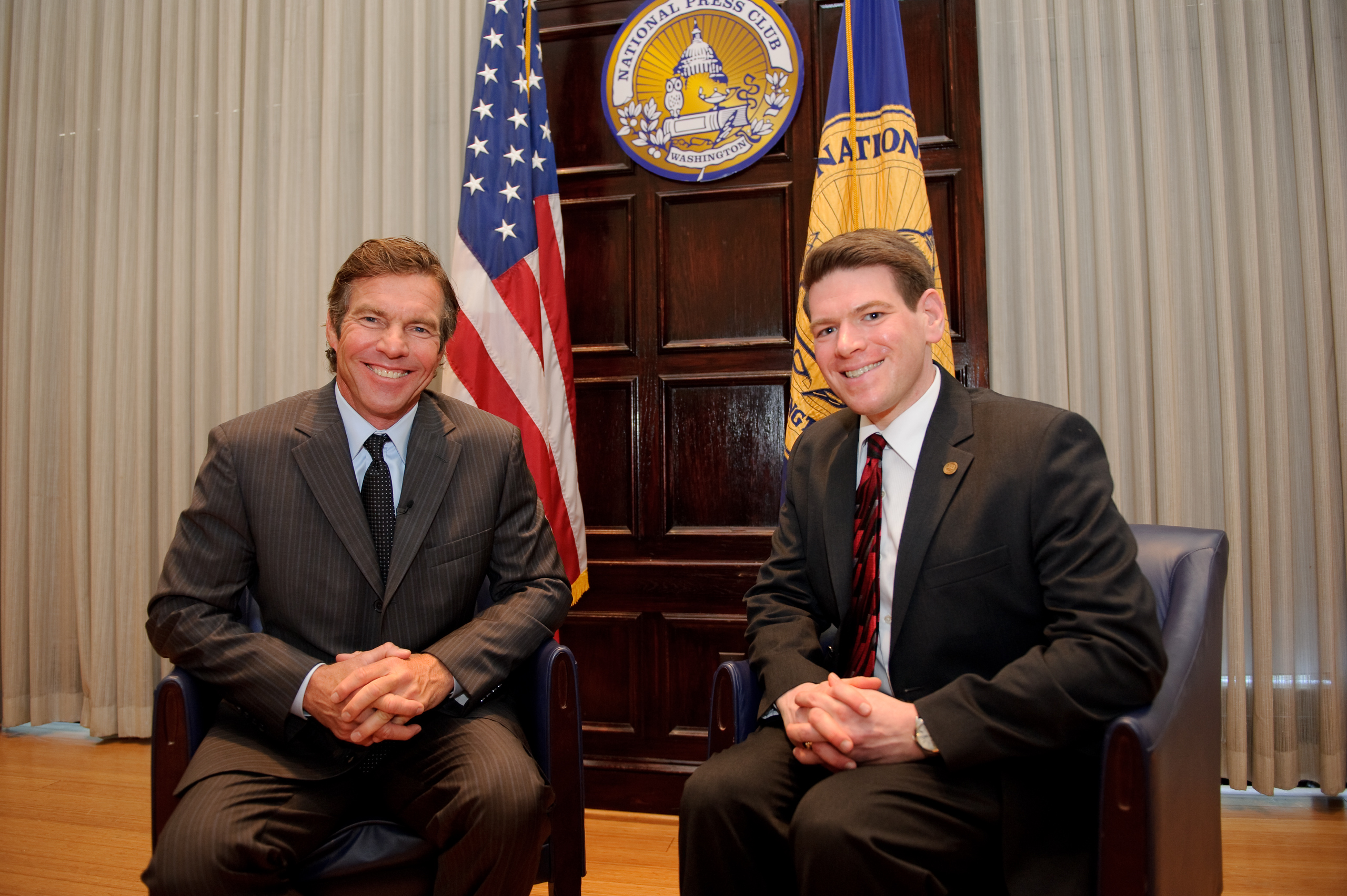 Two men in suits sit and smile for a photo in front of the U.S. flag and National Press Club banners in an official-looking setting.