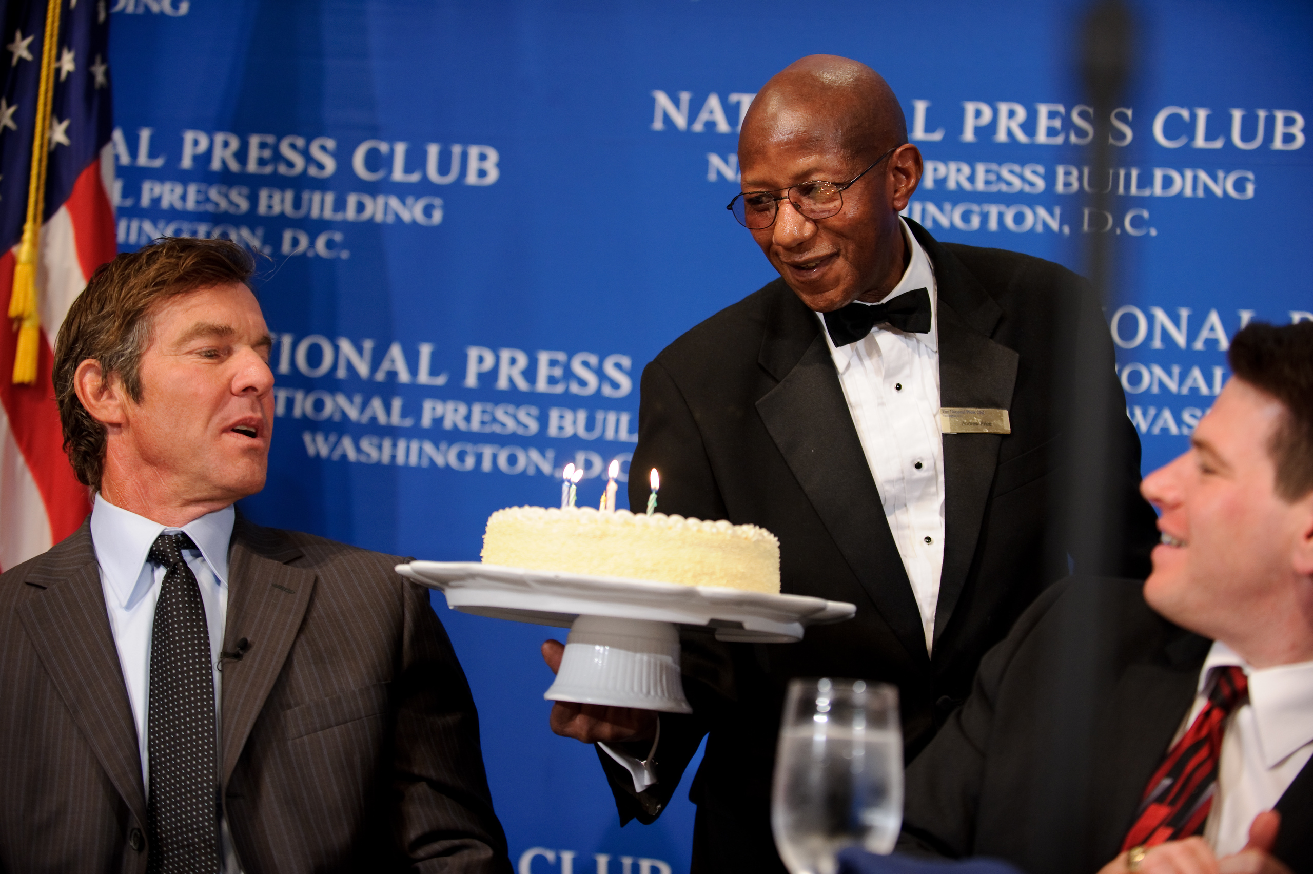 A server presents a cake with candles to a man in a suit at a National Press Club event, with another man seated nearby.