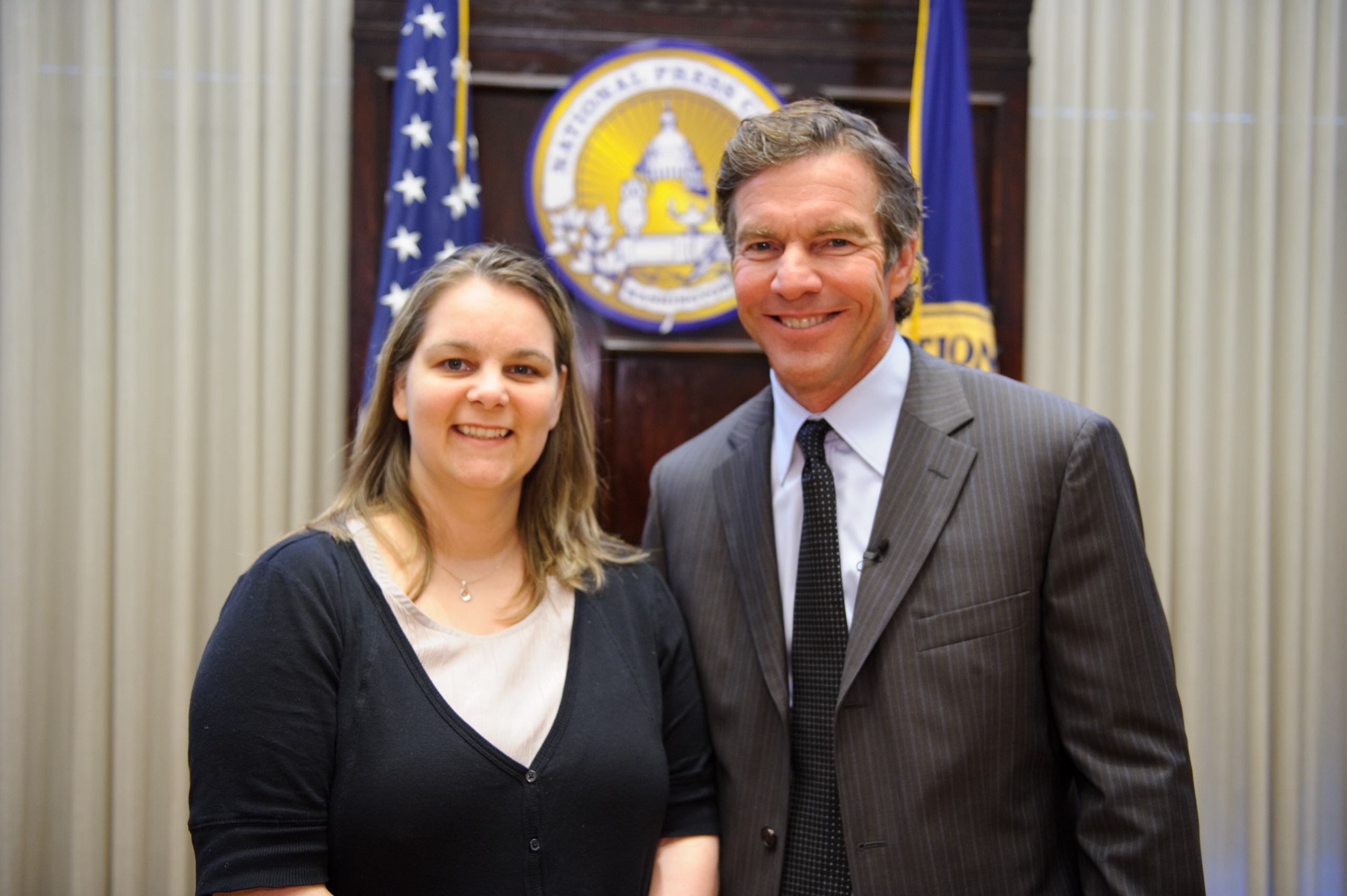 Two people stand smiling in front of American flags and an official seal in a formal indoor setting.