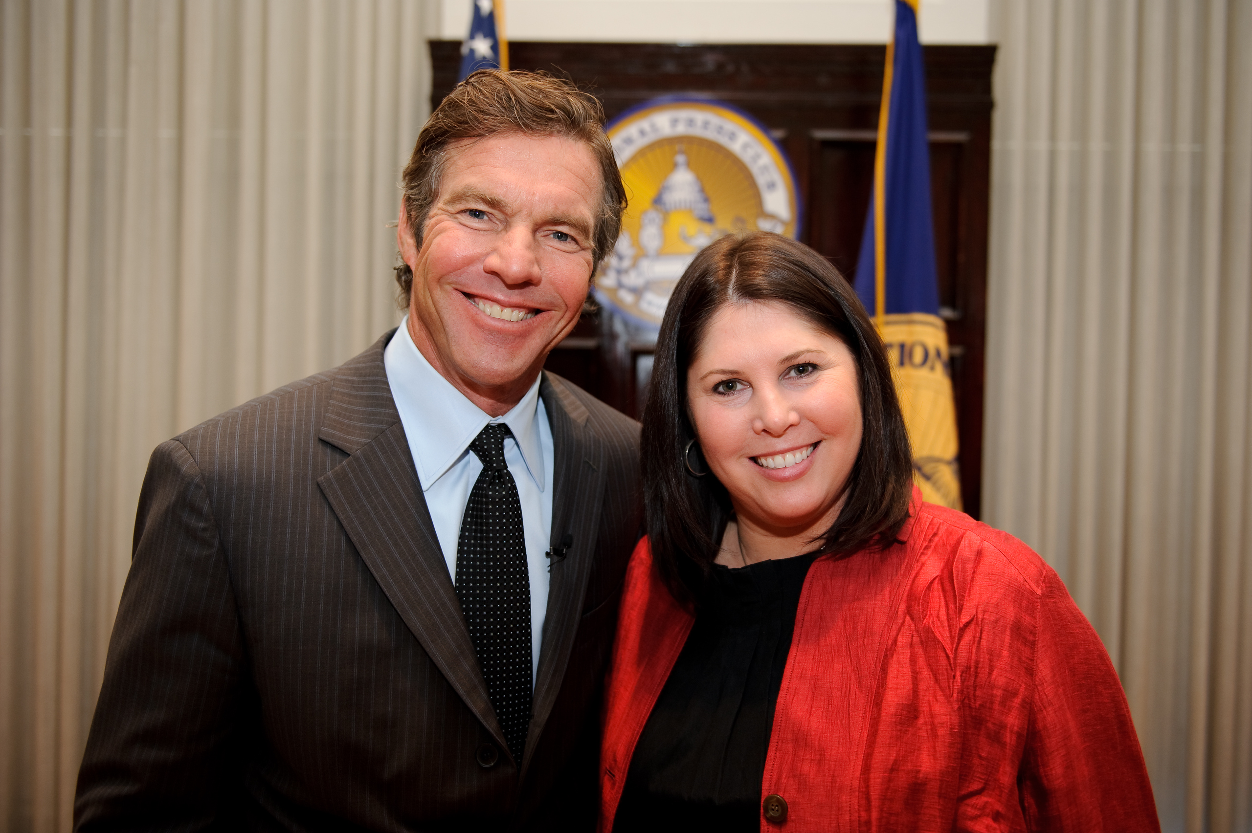 A man in a suit and a woman in a red jacket stand smiling together indoors, with flags and an official seal visible in the background.