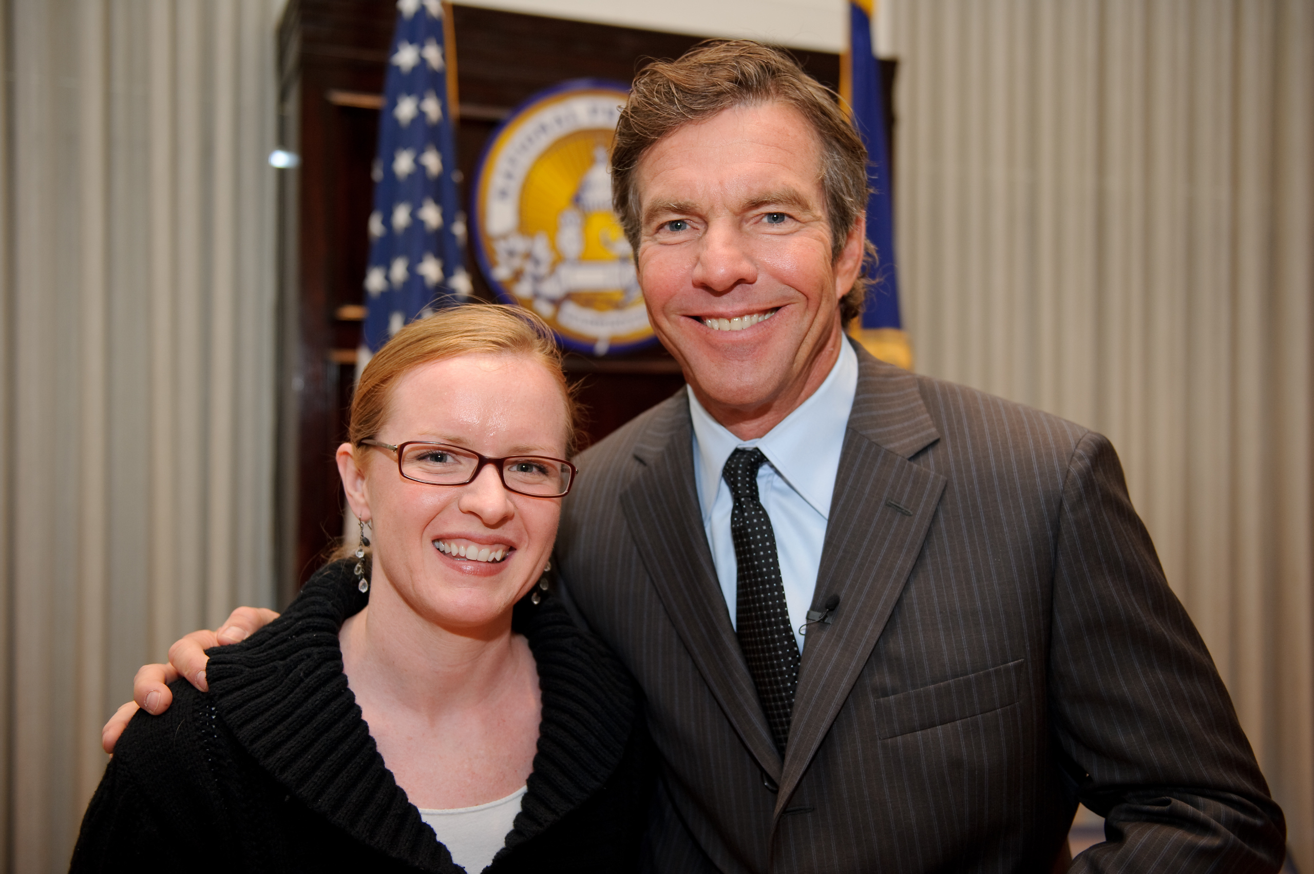 A woman and a man in a suit pose together, smiling, in front of an official seal and flags indoors.