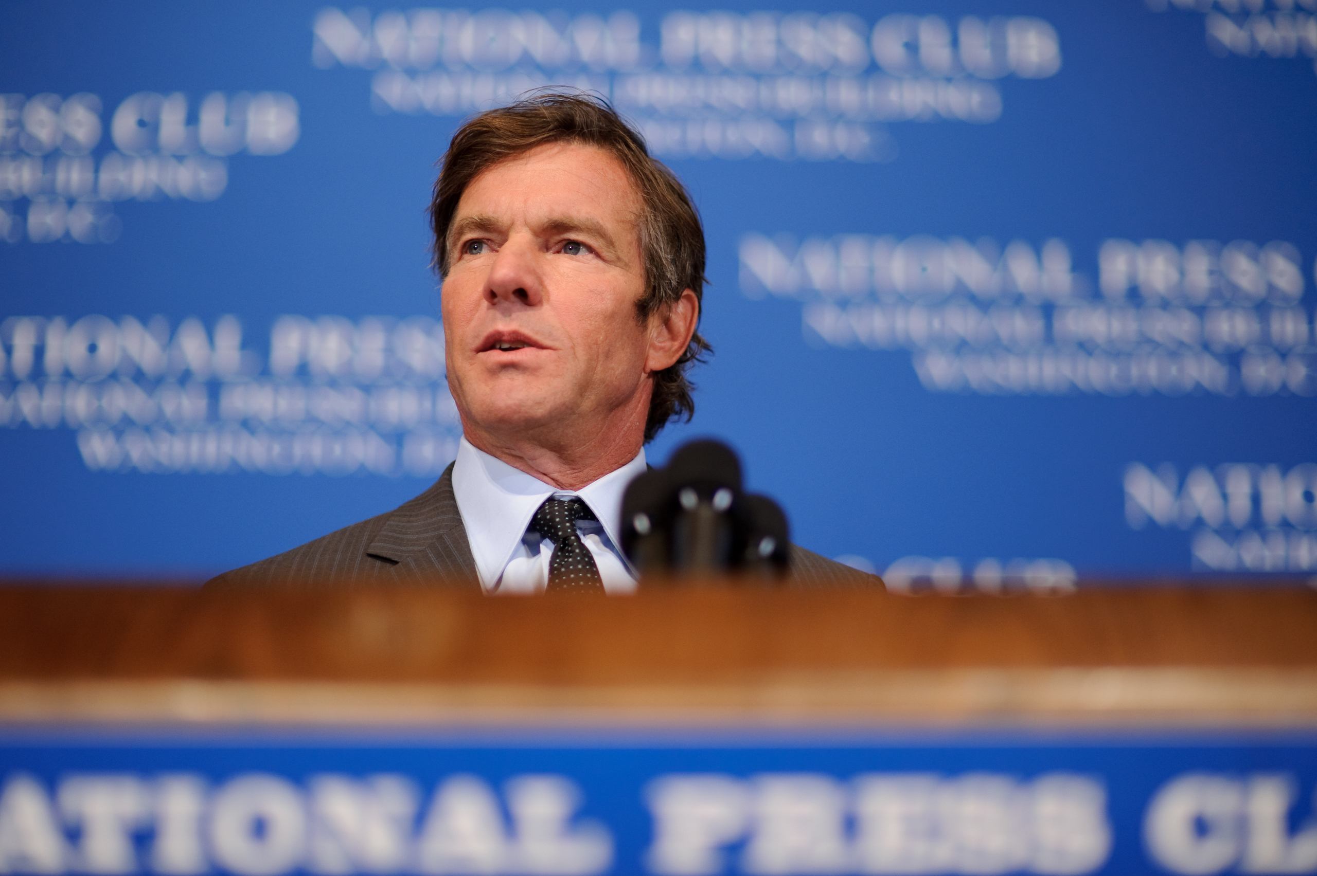 A man in a suit stands at a podium with microphones, speaking at a National Press Club event with a blue backdrop.
