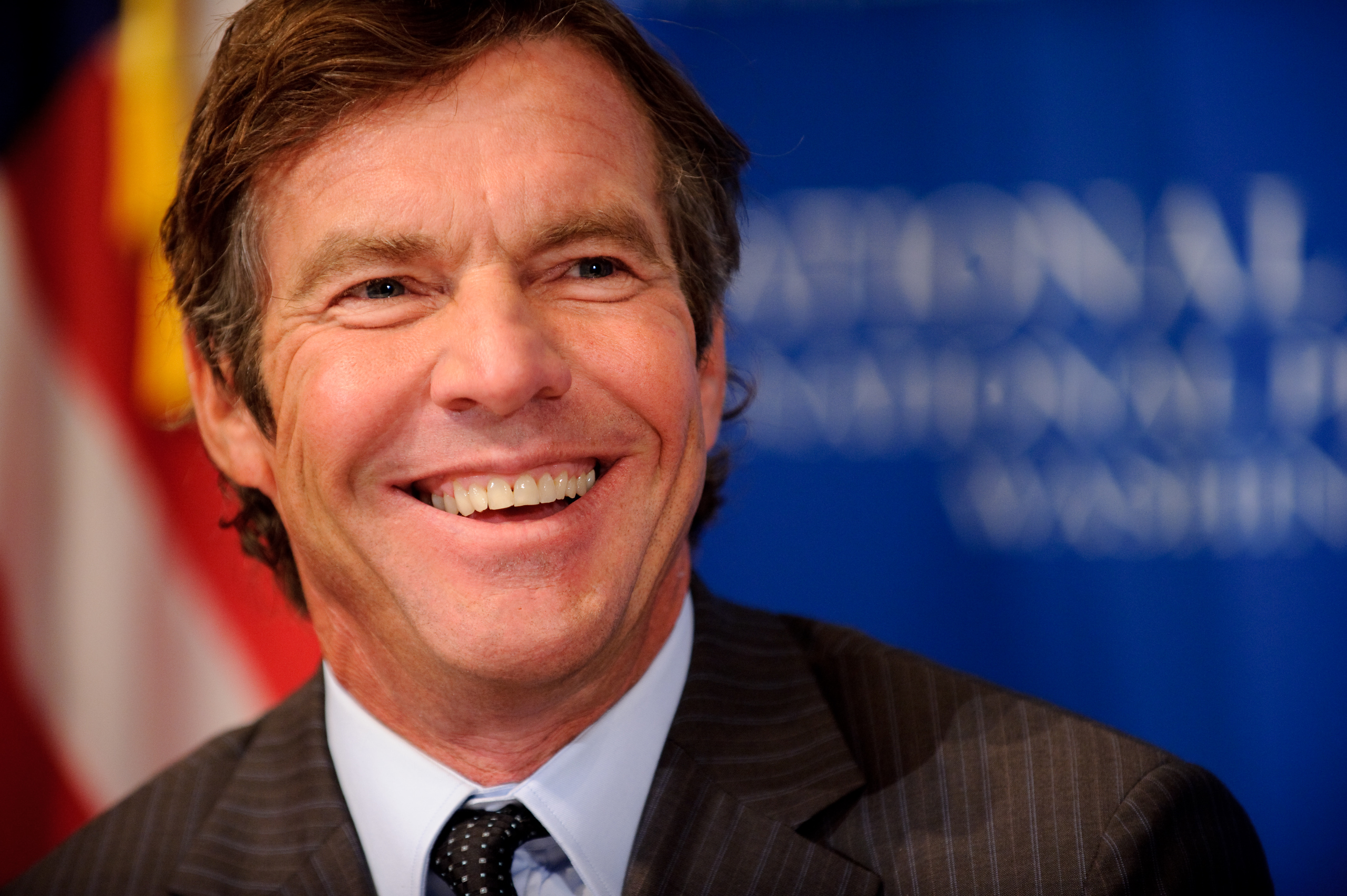 A man in a suit smiles in front of a blurred blue background with white text and an American flag.