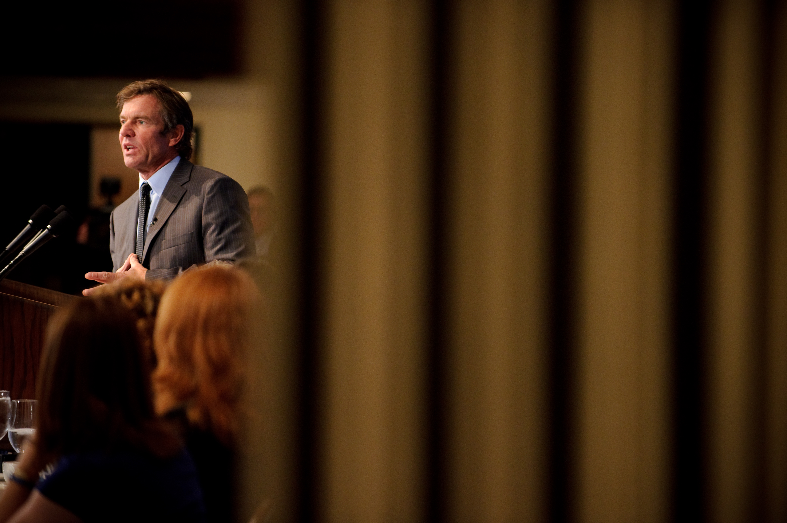 A man in a suit speaks at a podium to an audience; the view is partially obscured by vertical bars in the foreground.