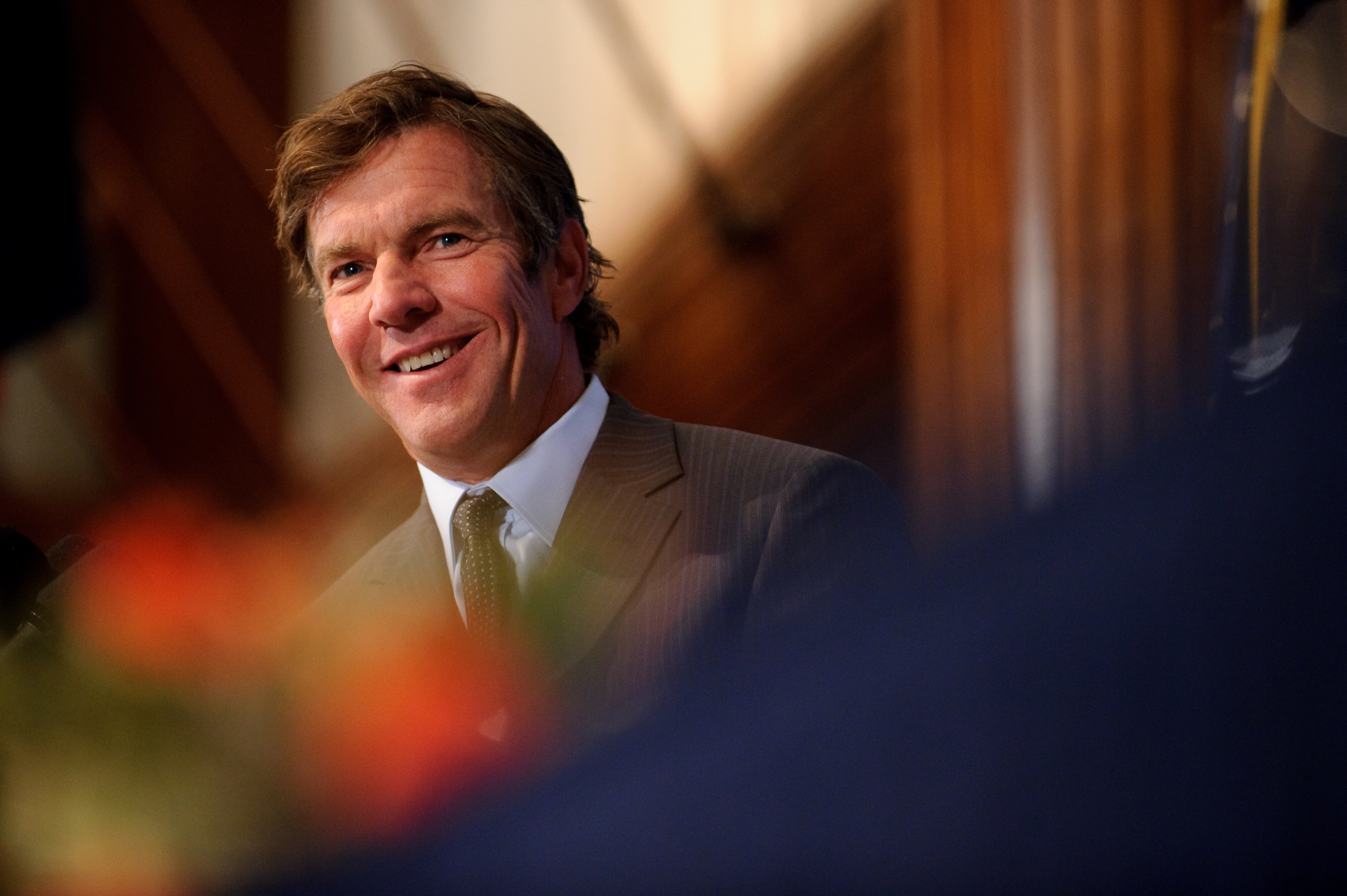 A man in a suit and tie is smiling while speaking at an indoor event, with blurred orange flowers in the foreground.