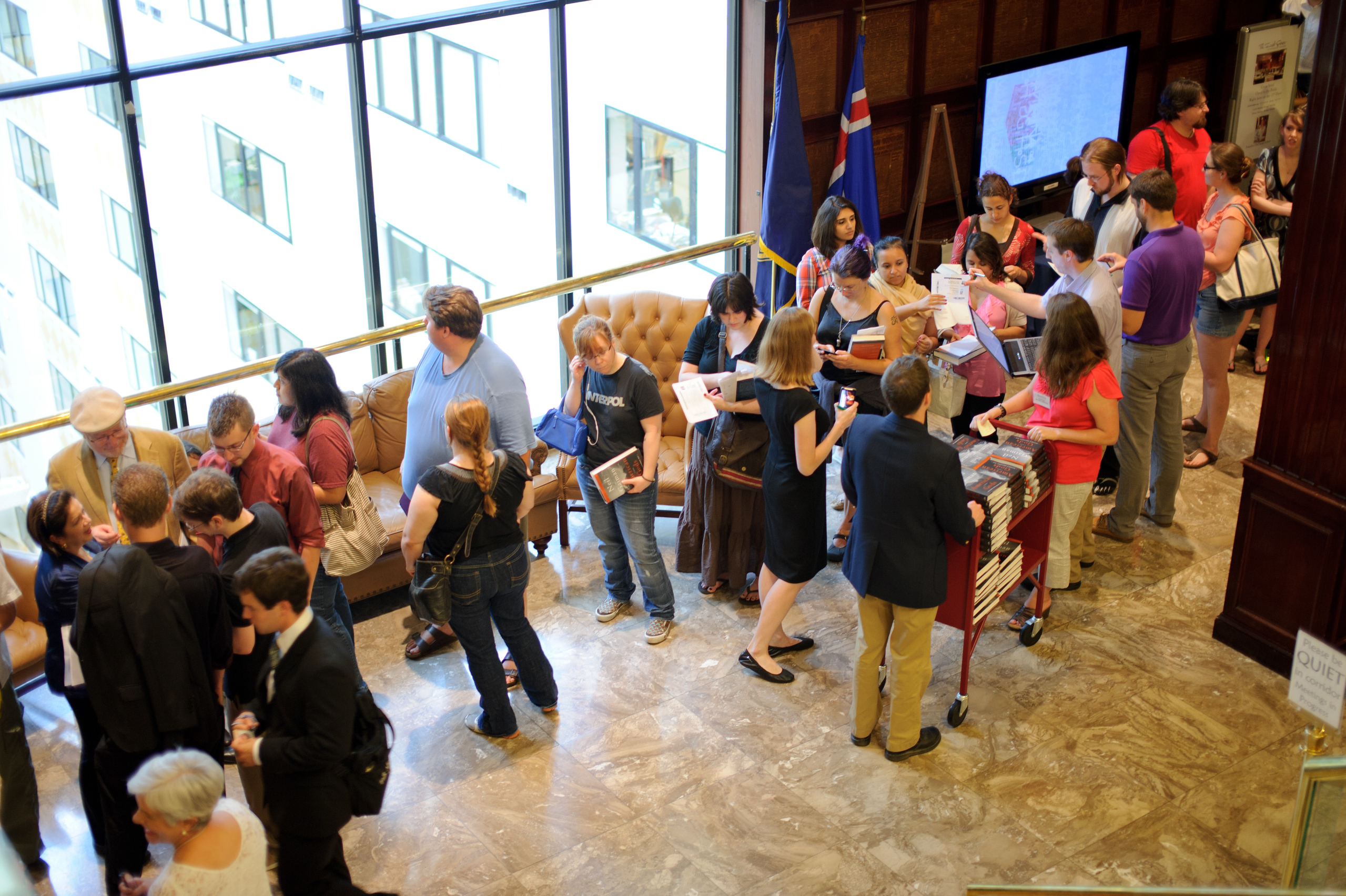 A group of people stand and interact in a lobby area near large windows; some are handing out or receiving papers near a display table.