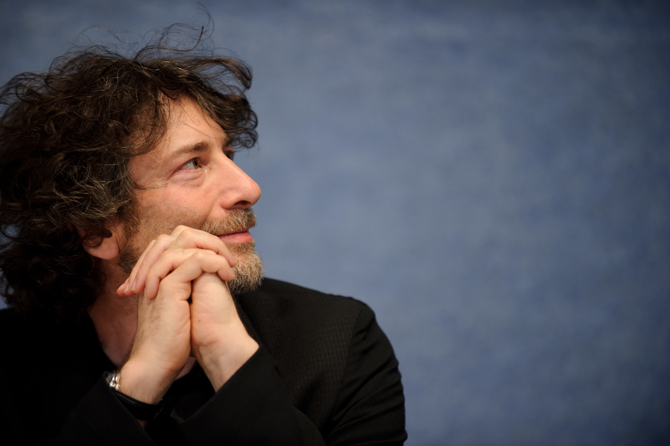 A man with curly dark hair and a beard sits with his hands clasped under his chin, looking to the left against a plain blue background.