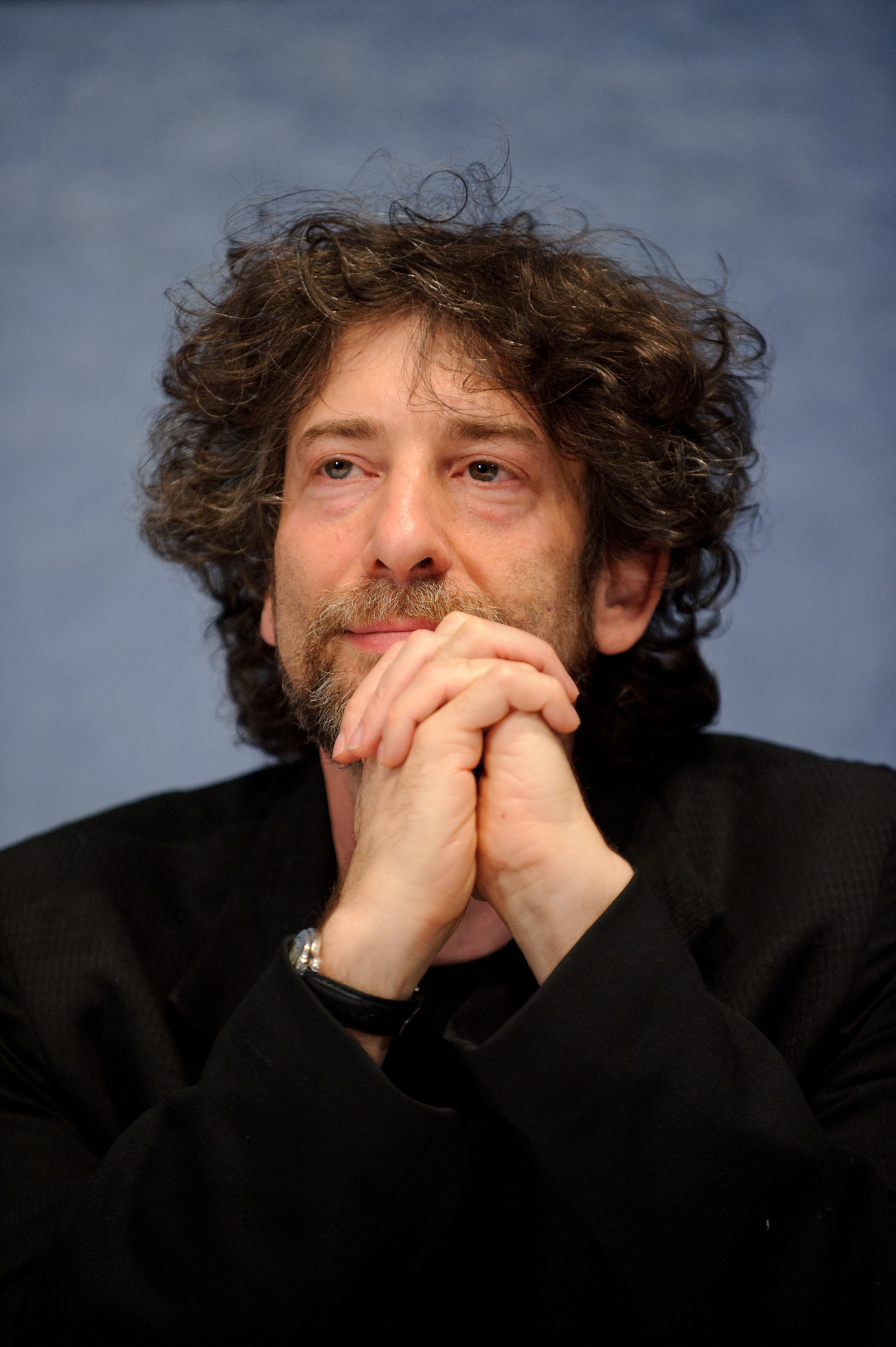 A man with curly hair and a beard rests his chin on his clasped hands, sitting in front of a blue background and wearing a black jacket.