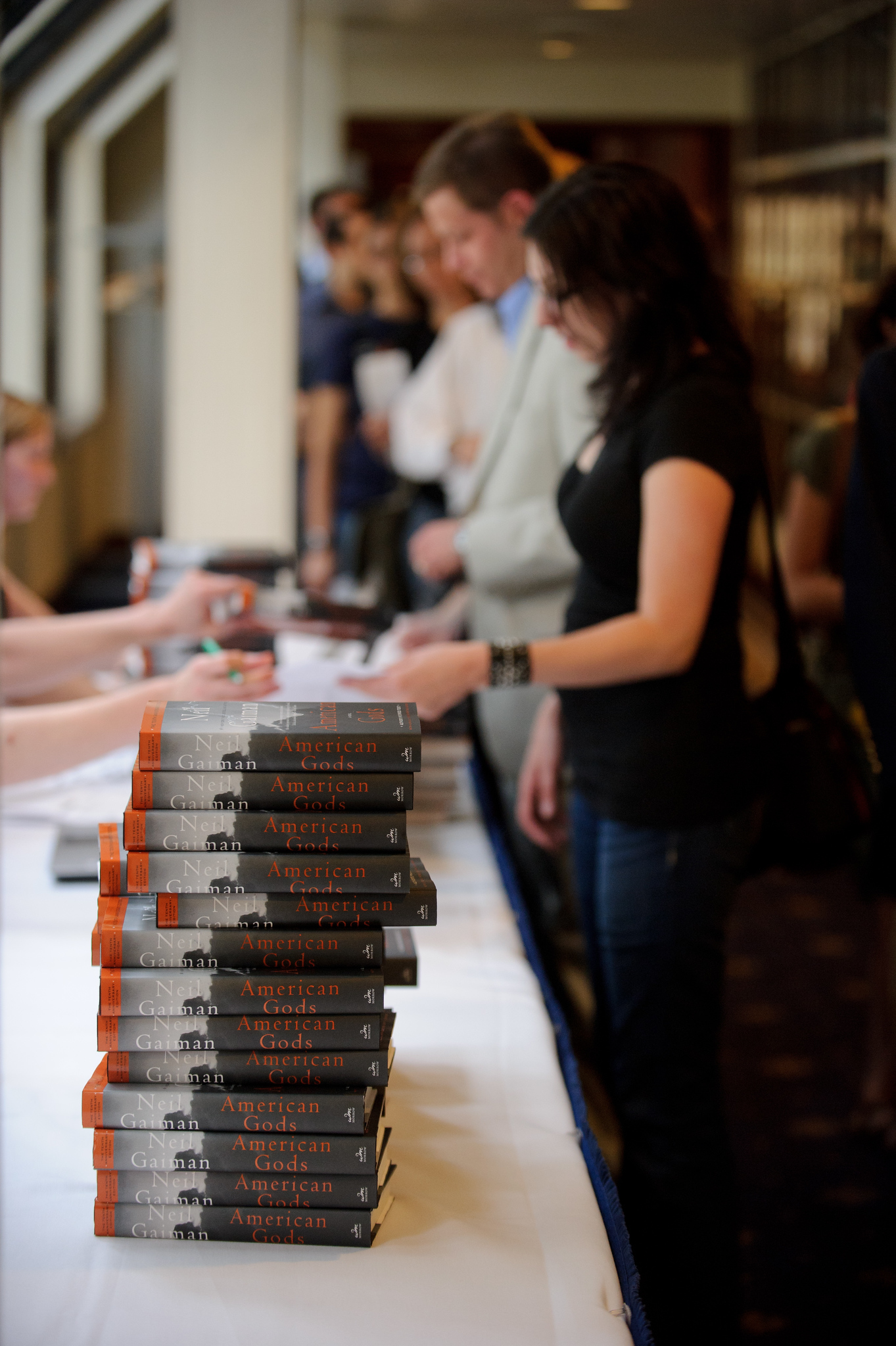 A stack of "American Gods" books sits on a table as people line up at a signing or registration event indoors.