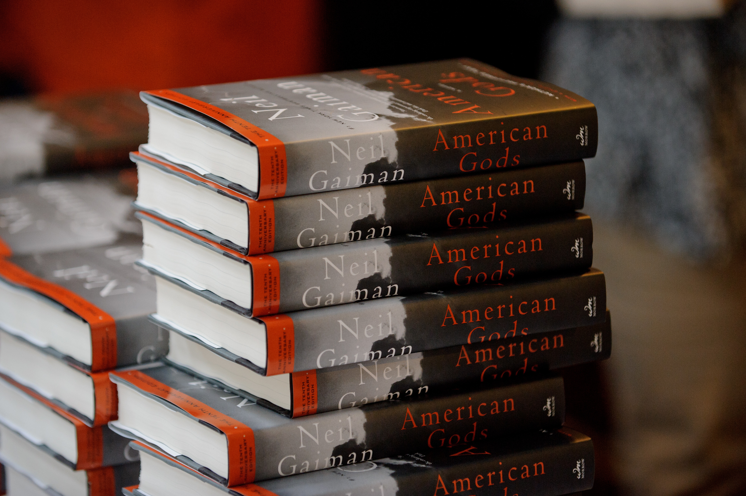 A stack of hardcover copies of the book "American Gods" by Neil Gaiman is shown, arranged neatly on a table.