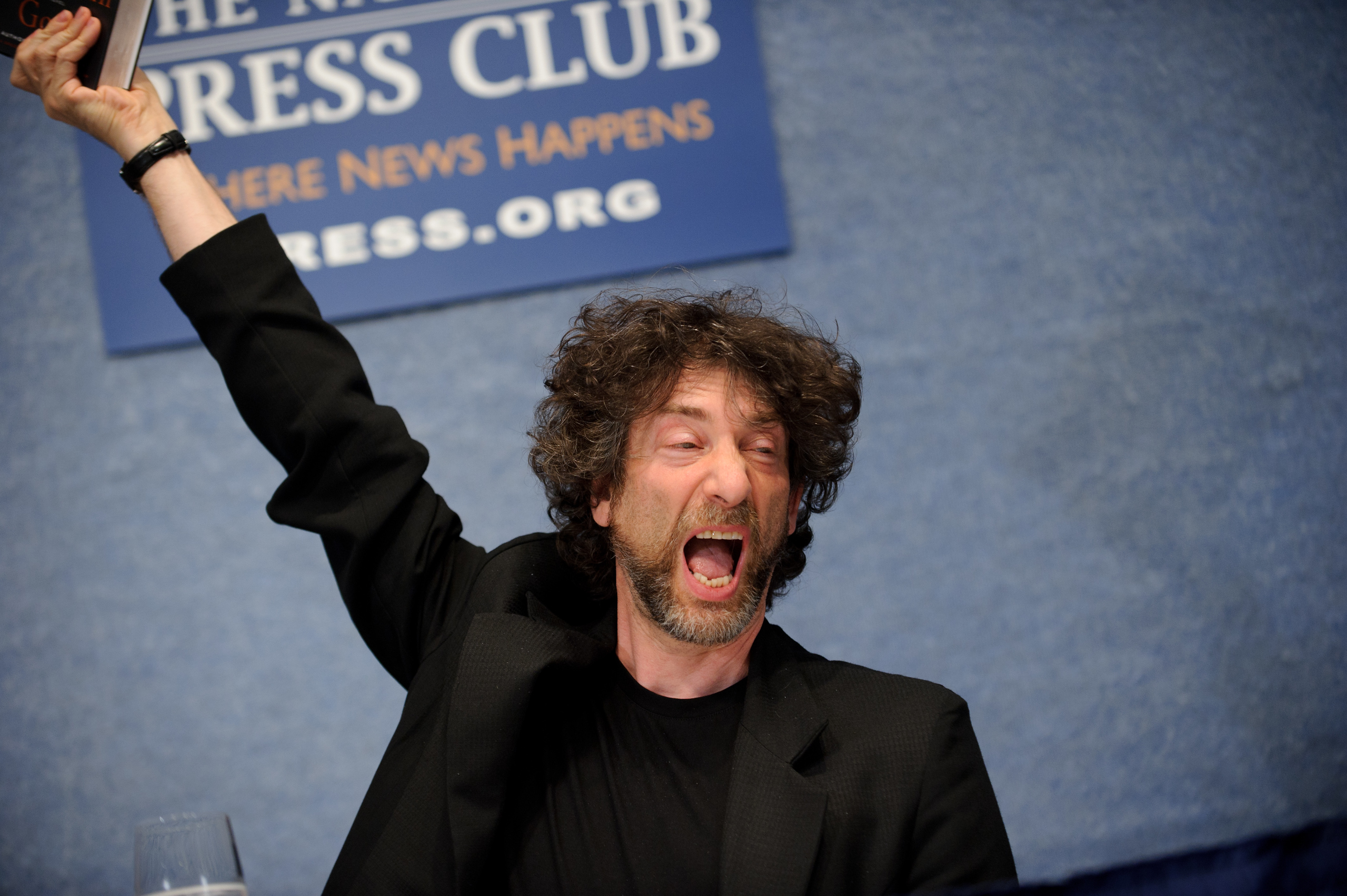A man with curly hair and a beard raises his arm and shouts enthusiastically while seated at a press conference.