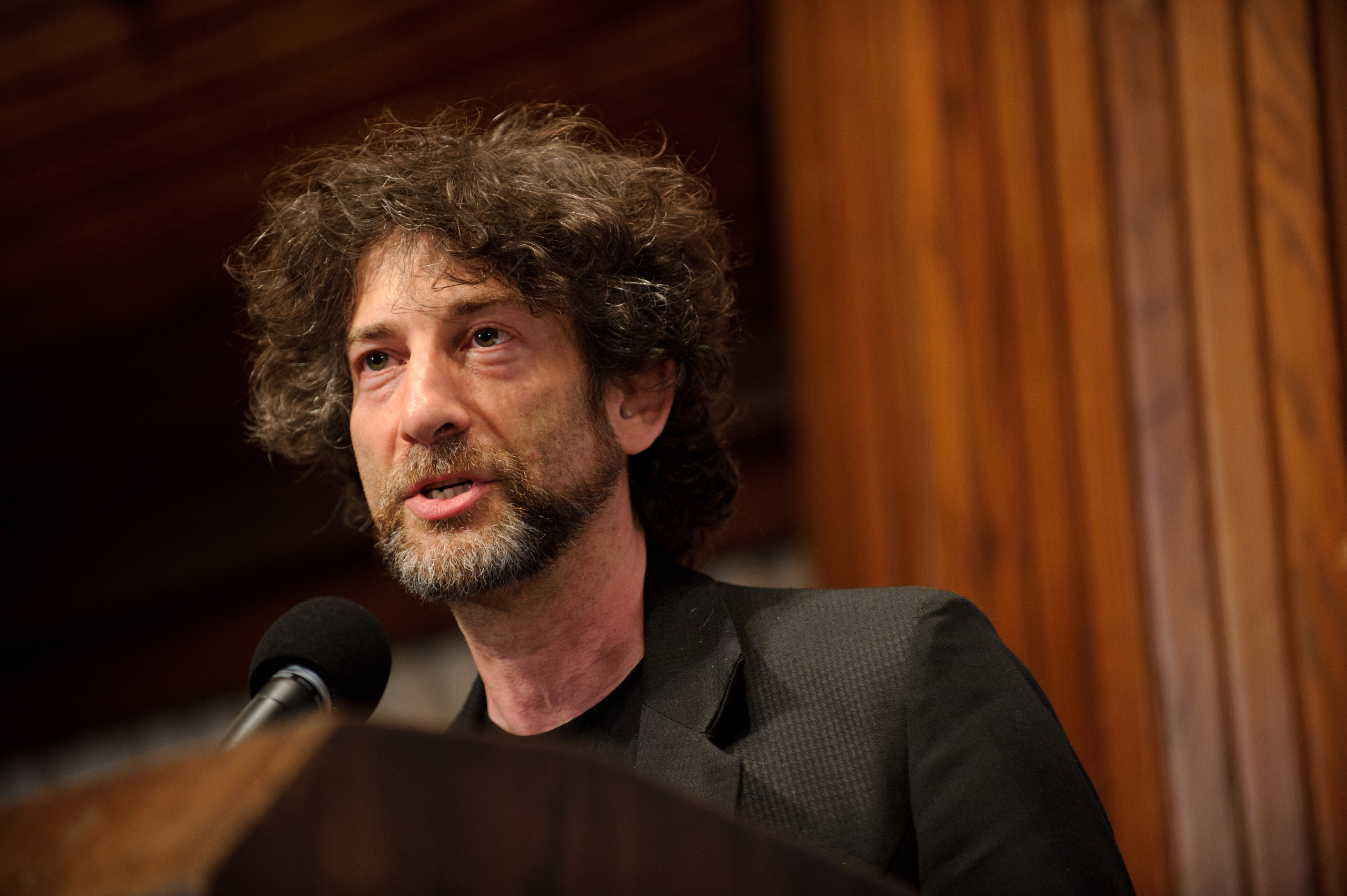 A man with curly hair and a beard speaks into a microphone at a podium, with wooden panels in the background.
