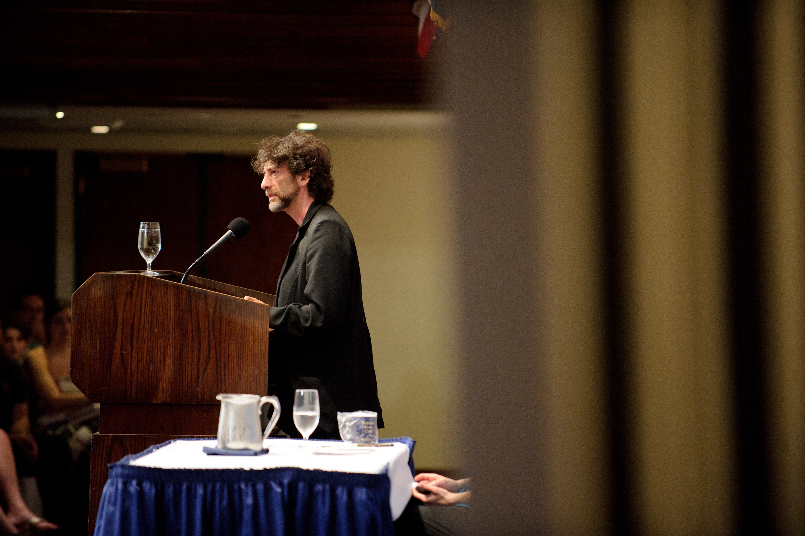 A man with curly hair speaks at a podium with a microphone, while an audience listens. A table with glasses and a pitcher is visible in the foreground.