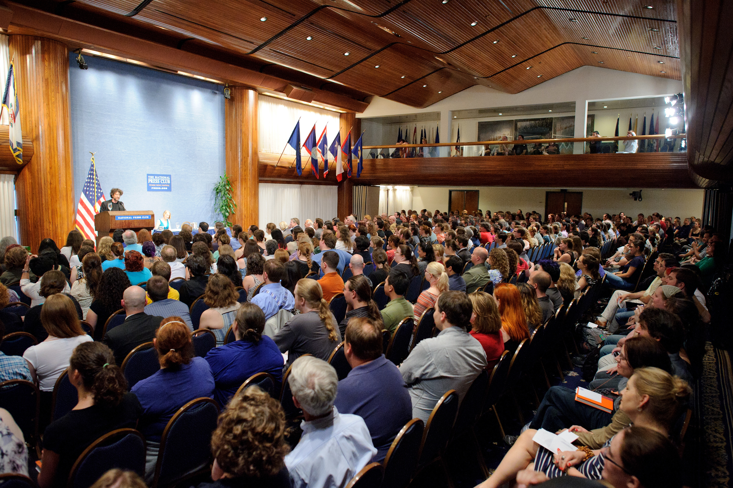 A woman stands at a podium addressing a large seated audience in a wood-paneled conference hall with flags displayed on the walls.