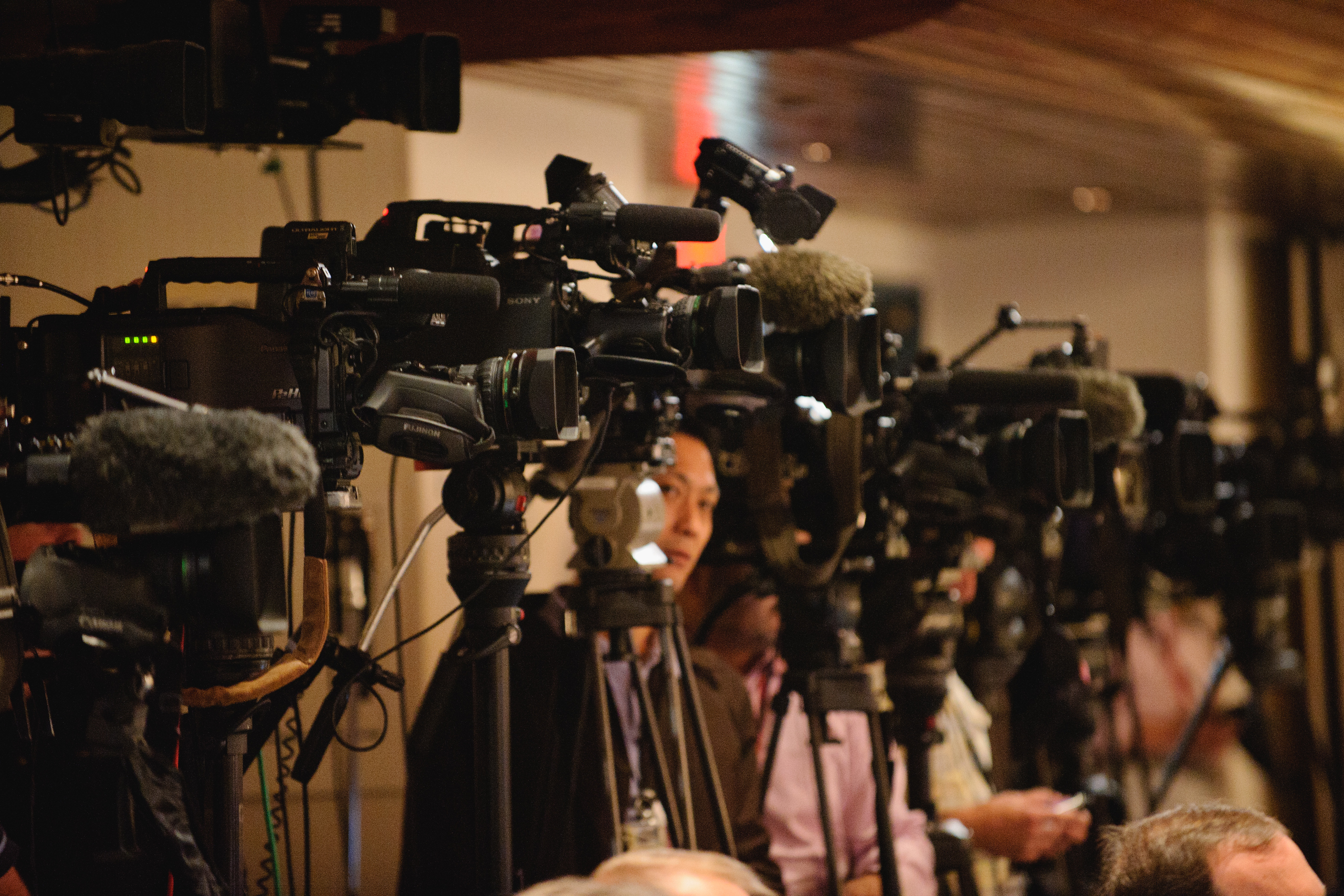 A row of professional video cameras and microphones set up on tripods, focused in the same direction, with people standing among the equipment indoors.