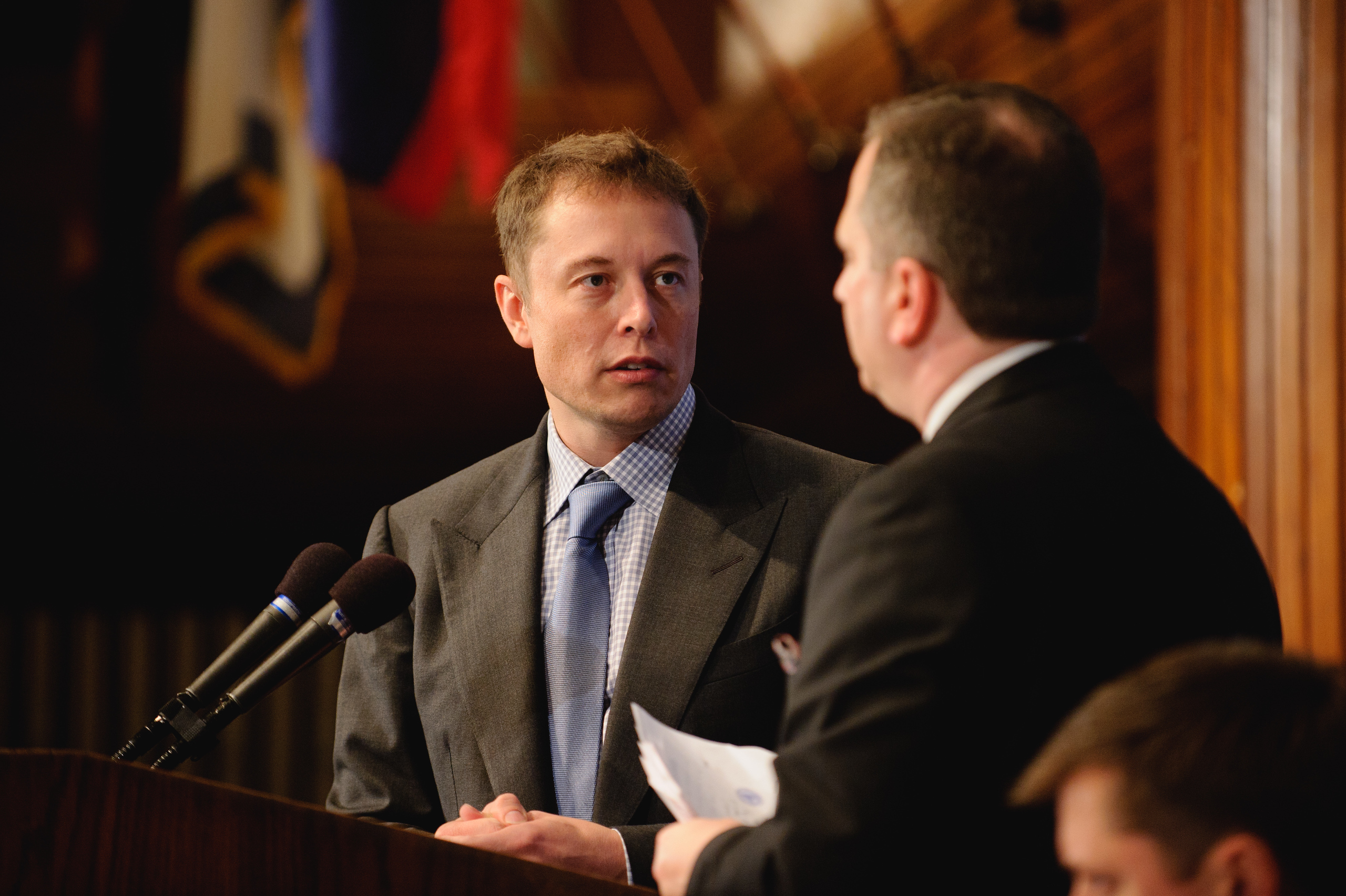 Two men in suits have a conversation at a podium, with microphones in front of them and flags visible in the background.