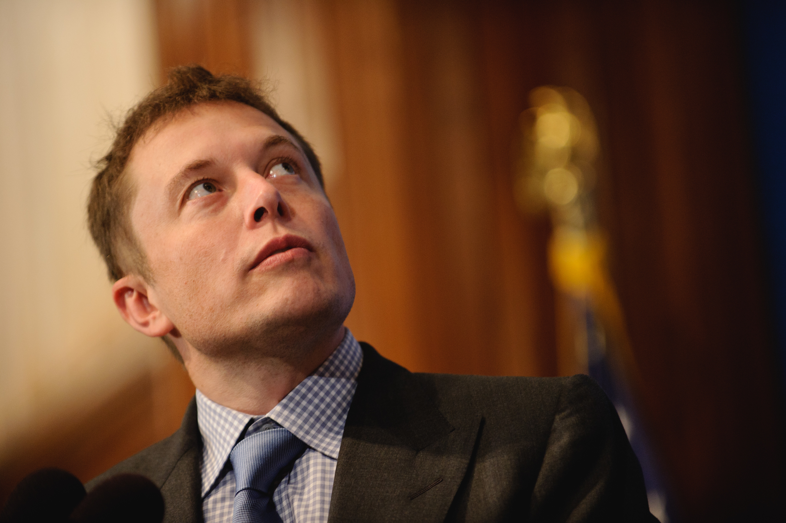 A man in a suit and blue tie looks upward, standing indoors with blurred background elements including a flag.