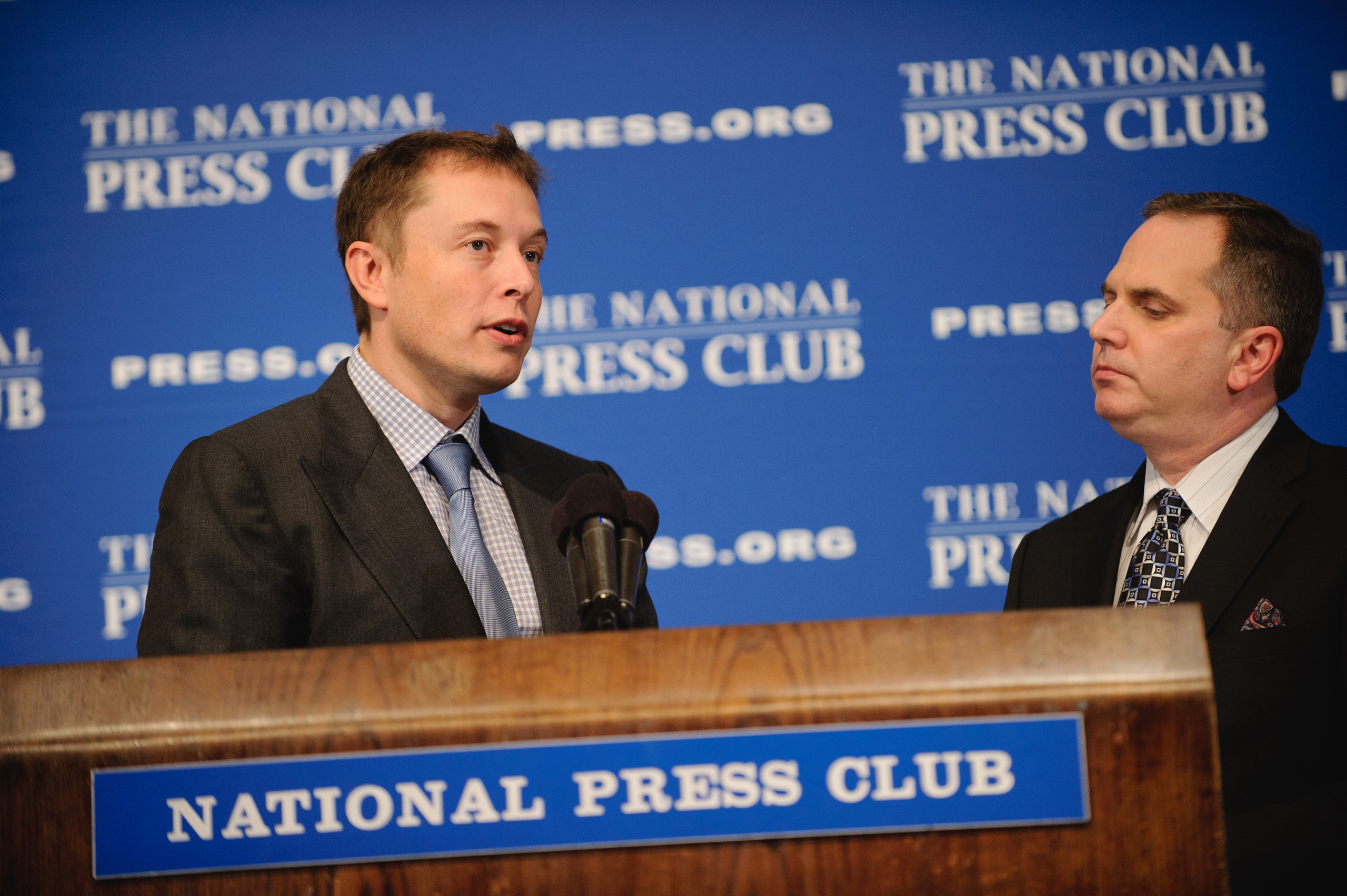 Two men in suits speak at a podium labeled "National Press Club" with a blue backdrop displaying "The National Press Club" and its website.