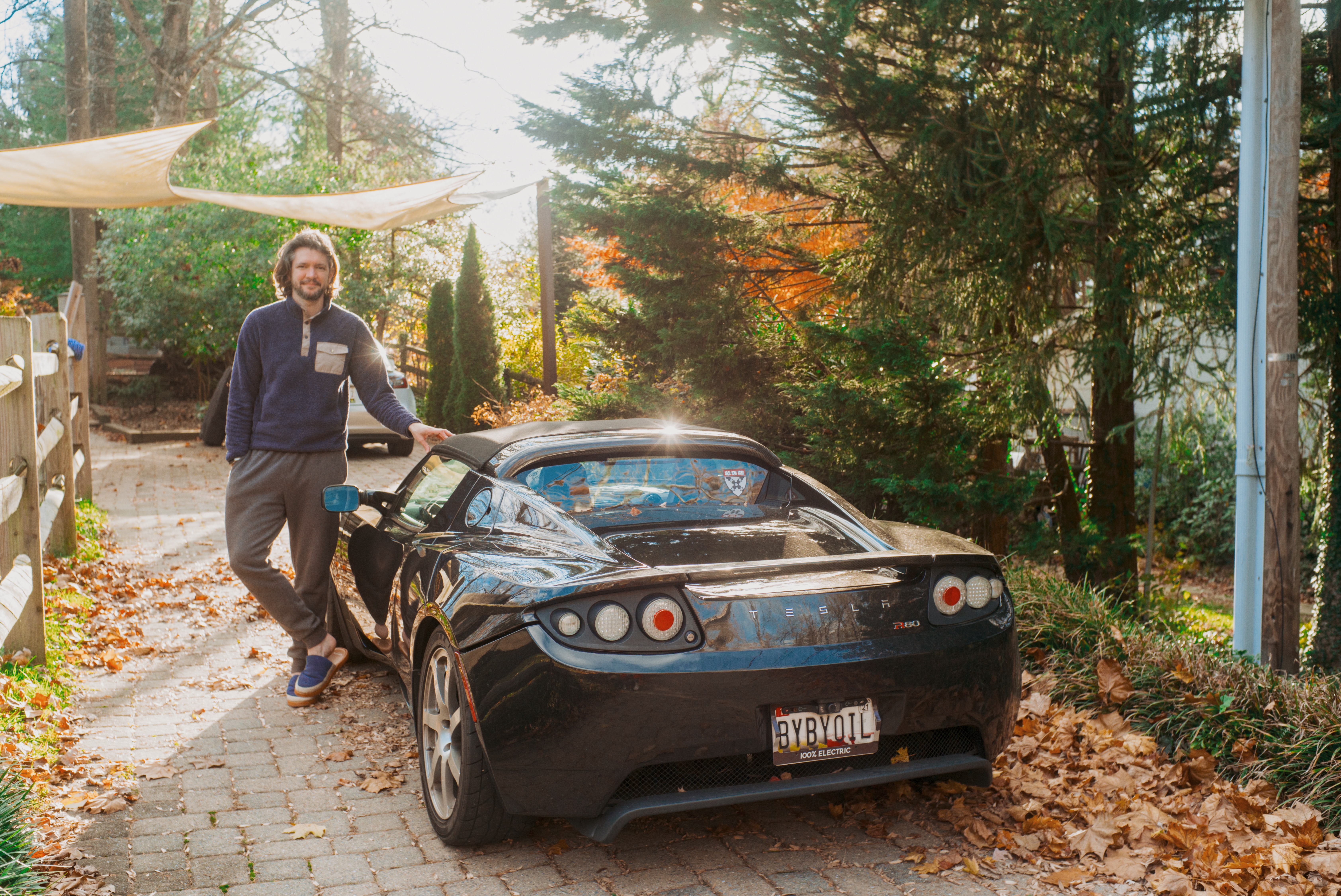 A person stands next to a black convertible sports car parked on a leaf-covered driveway surrounded by trees and autumn foliage.
