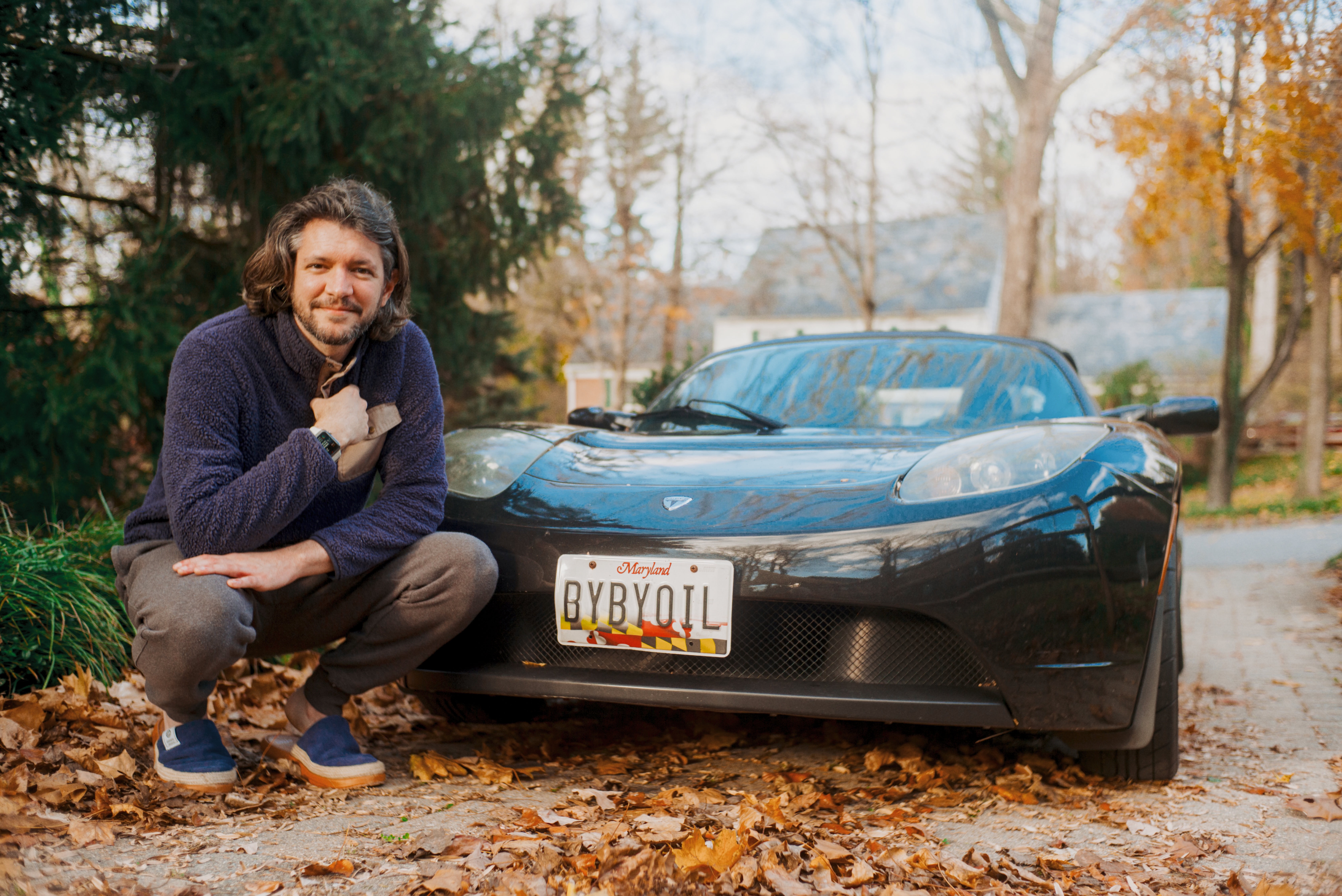 A man crouches next to a black Tesla Roadster with an Ohio license plate reading "BYBYOIL" on a leaf-covered driveway in a suburban neighborhood.