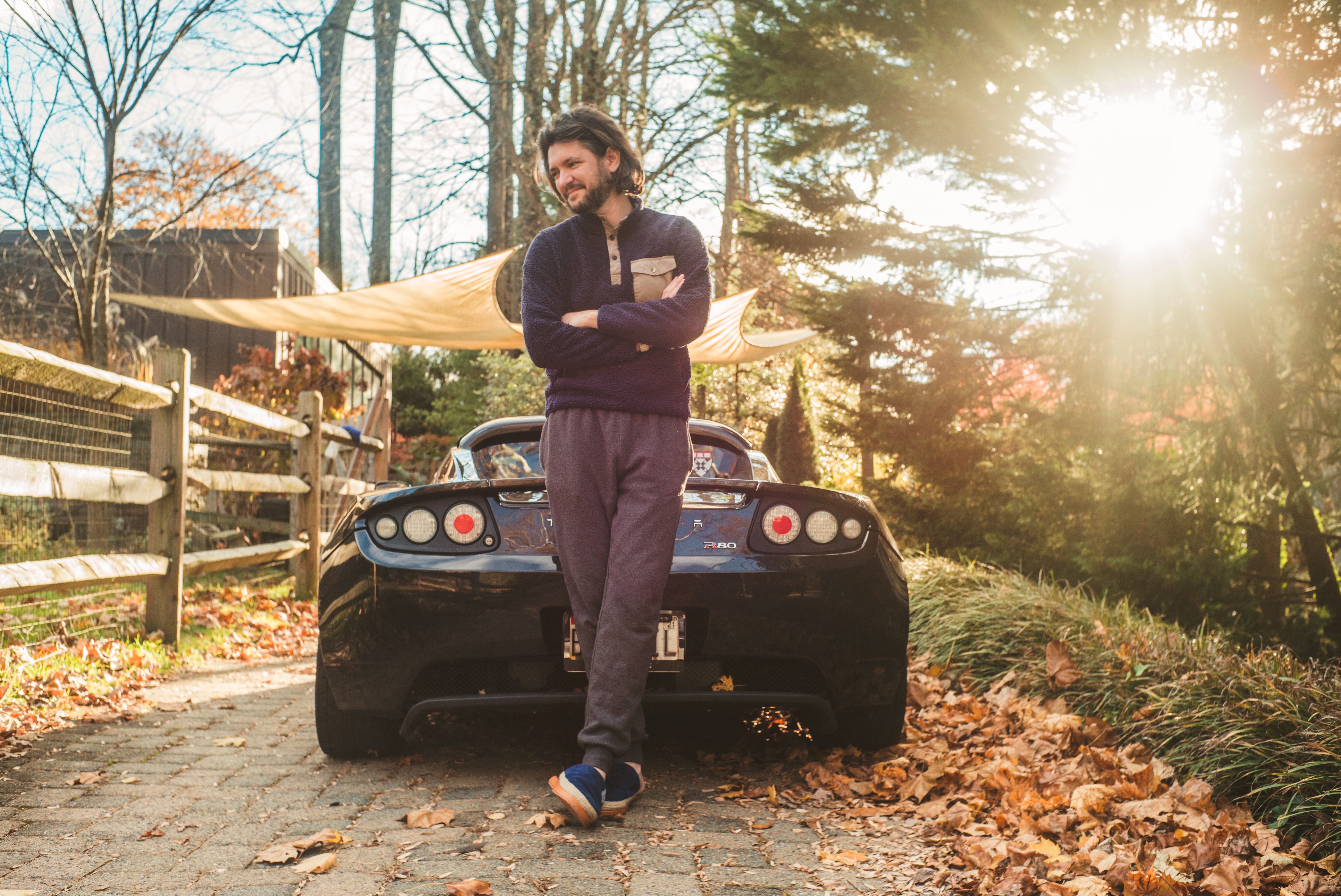 A man stands with crossed arms in front of a parked sports car on a leaf-covered driveway, with trees and sunlight in the background.