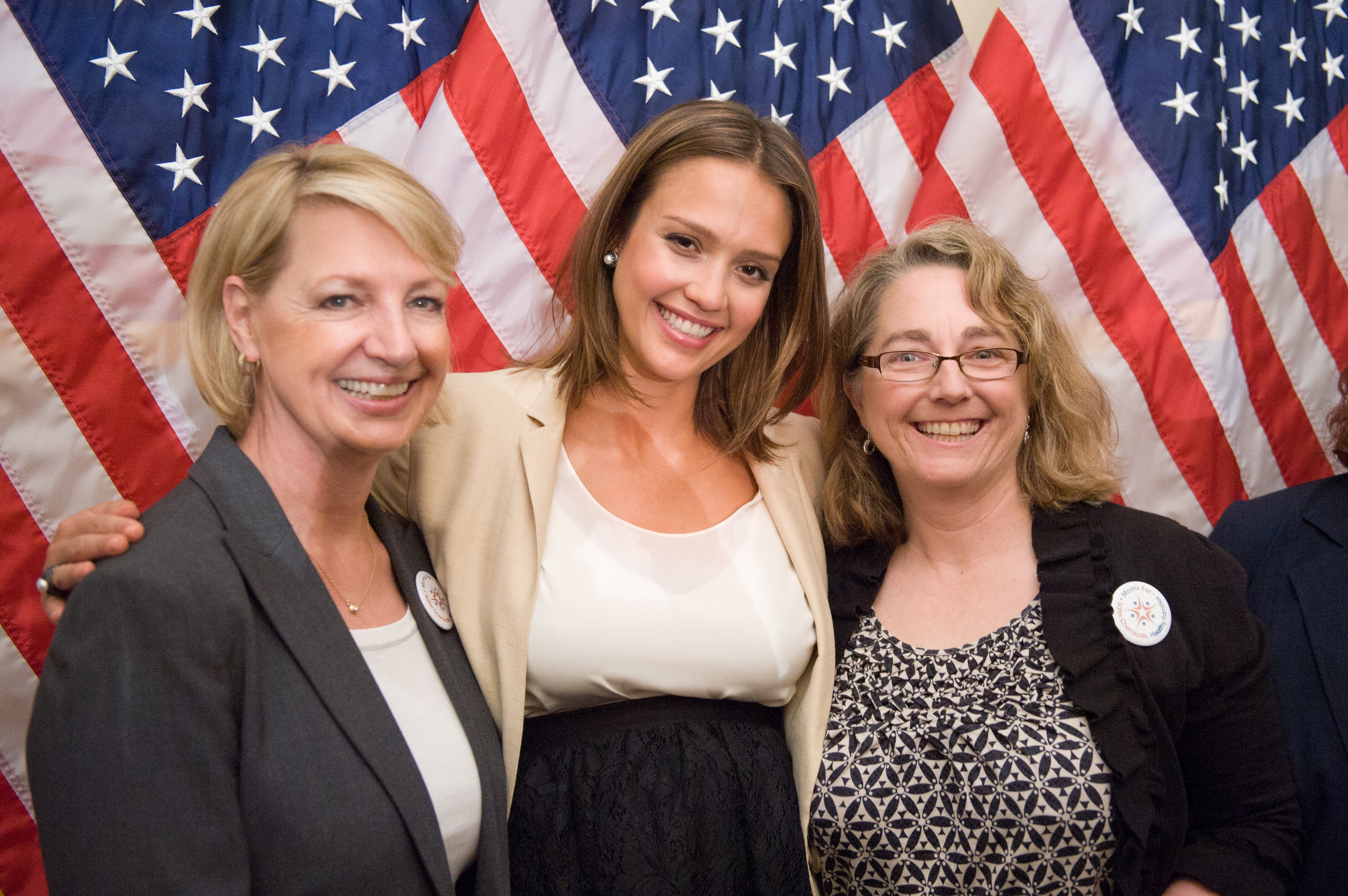 Three women stand smiling in front of multiple American flags, wearing business or semi-formal attire.