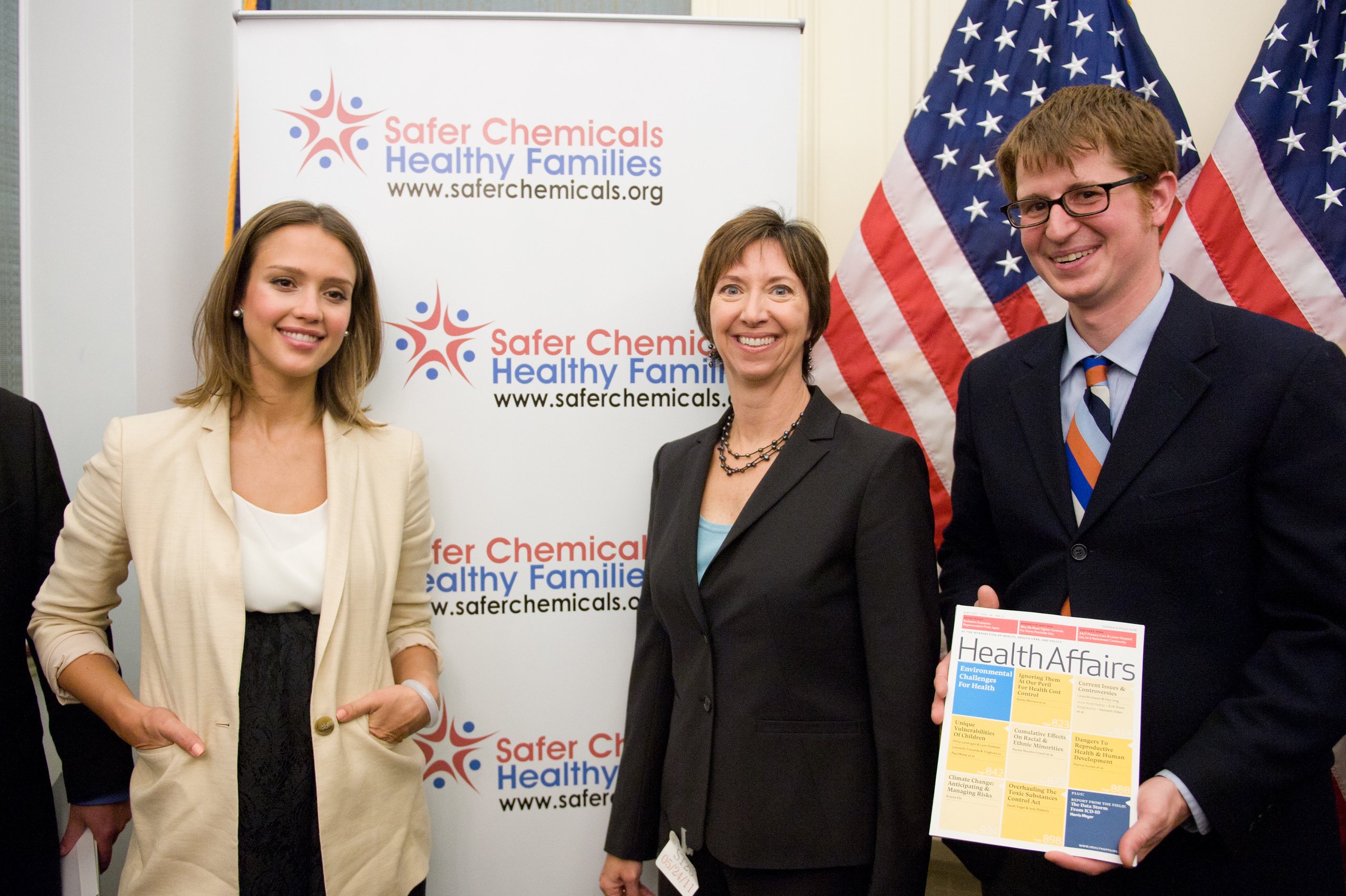 Three people stand in front of a "Safer Chemicals Healthy Families" banner and U.S. flags; one holds a "Health Affairs" magazine.