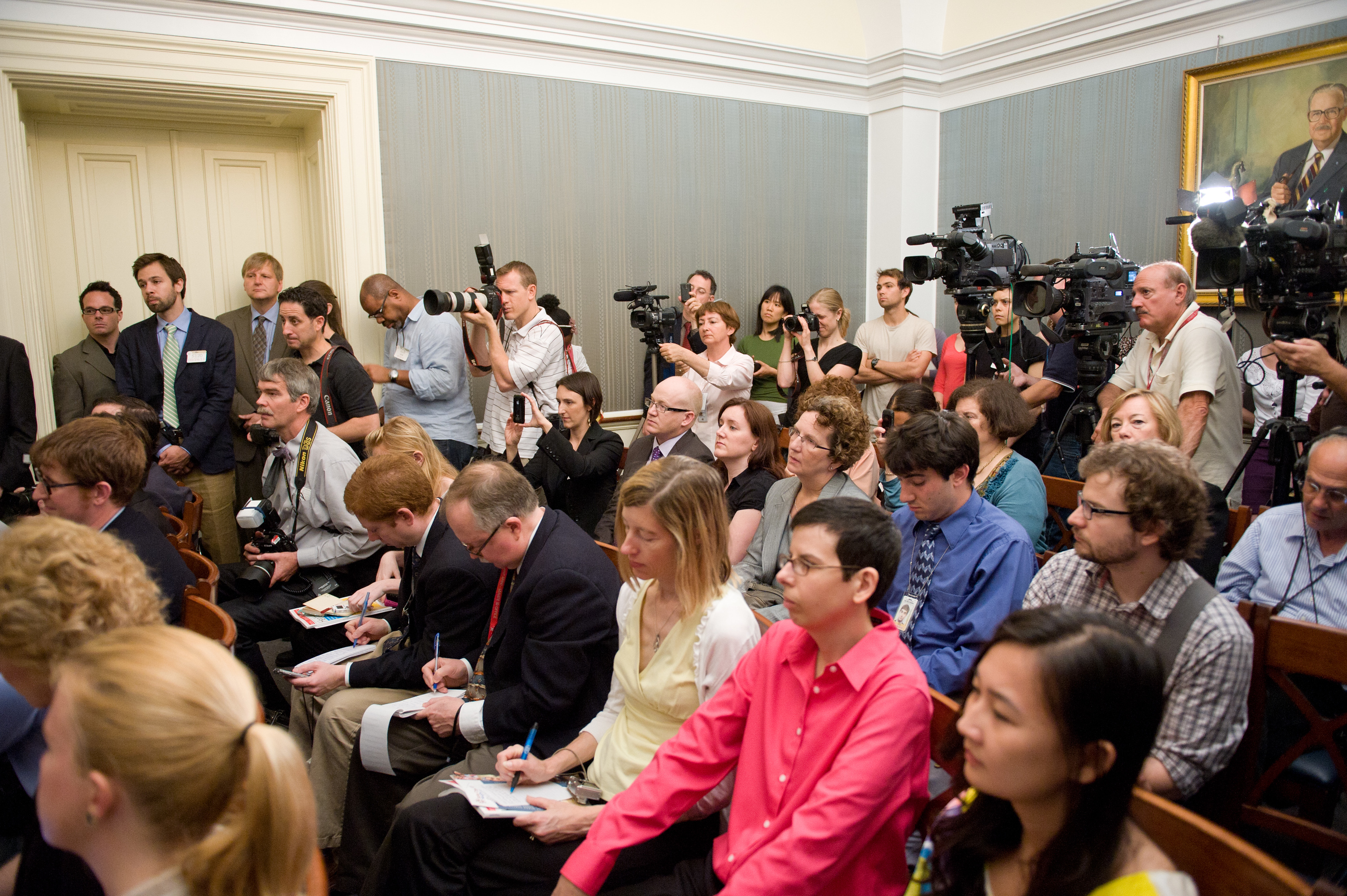 A group of journalists and photographers with cameras and notepads attend a press conference in a room with a portrait on the wall.
