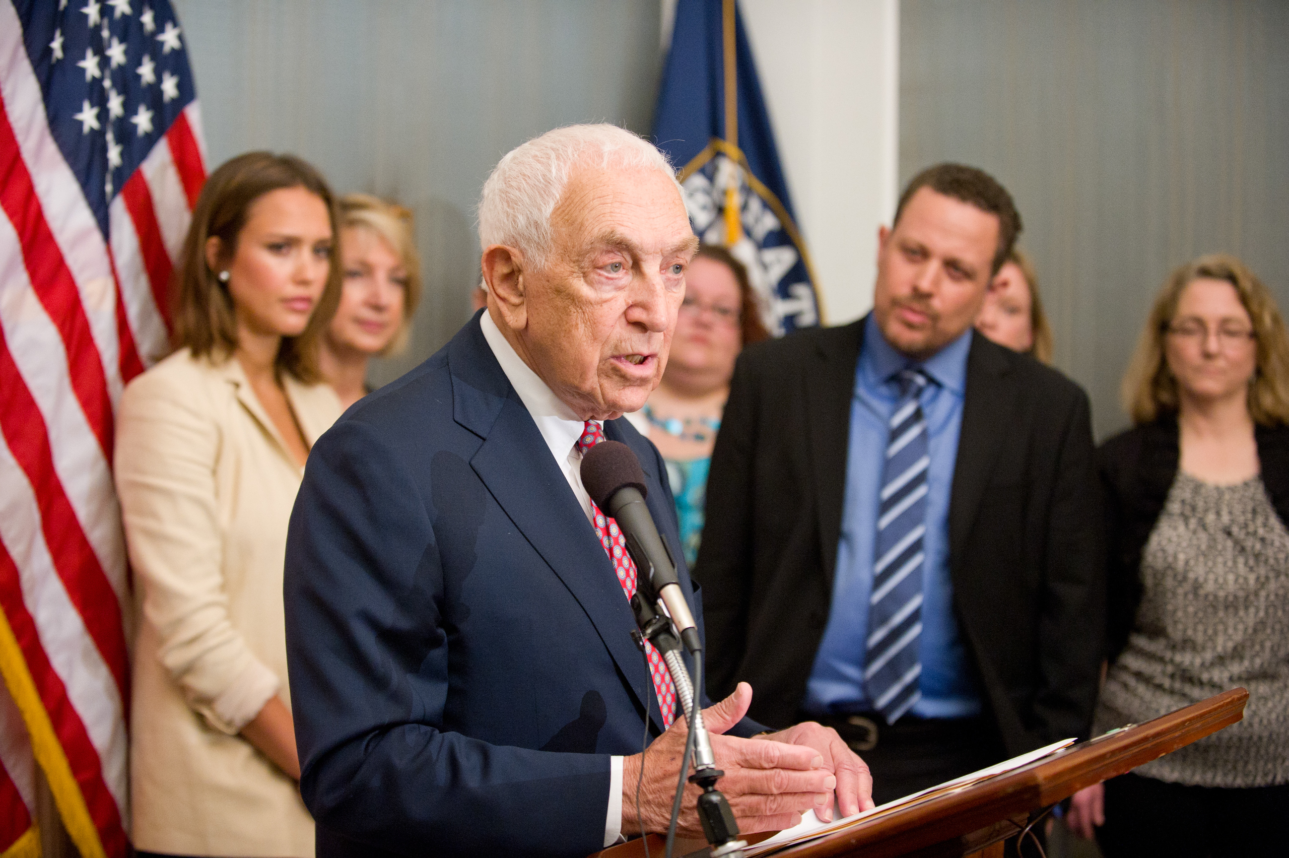 An elderly man in a suit speaks at a podium with a microphone, while several people stand behind him near American flags and a blue flag.