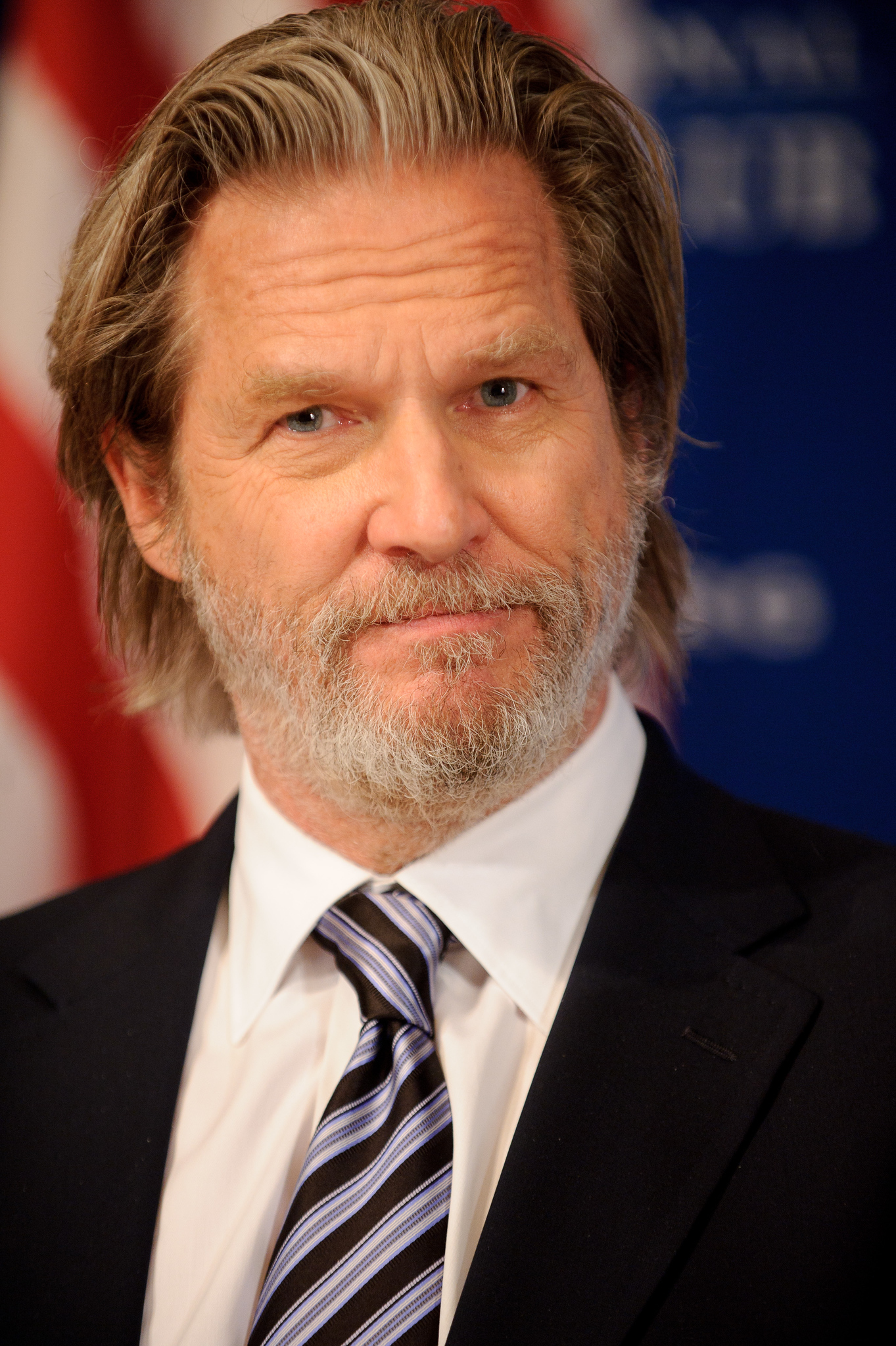 A man with gray hair and a beard is wearing a suit and striped tie, standing in front of an American flag and a blue background.