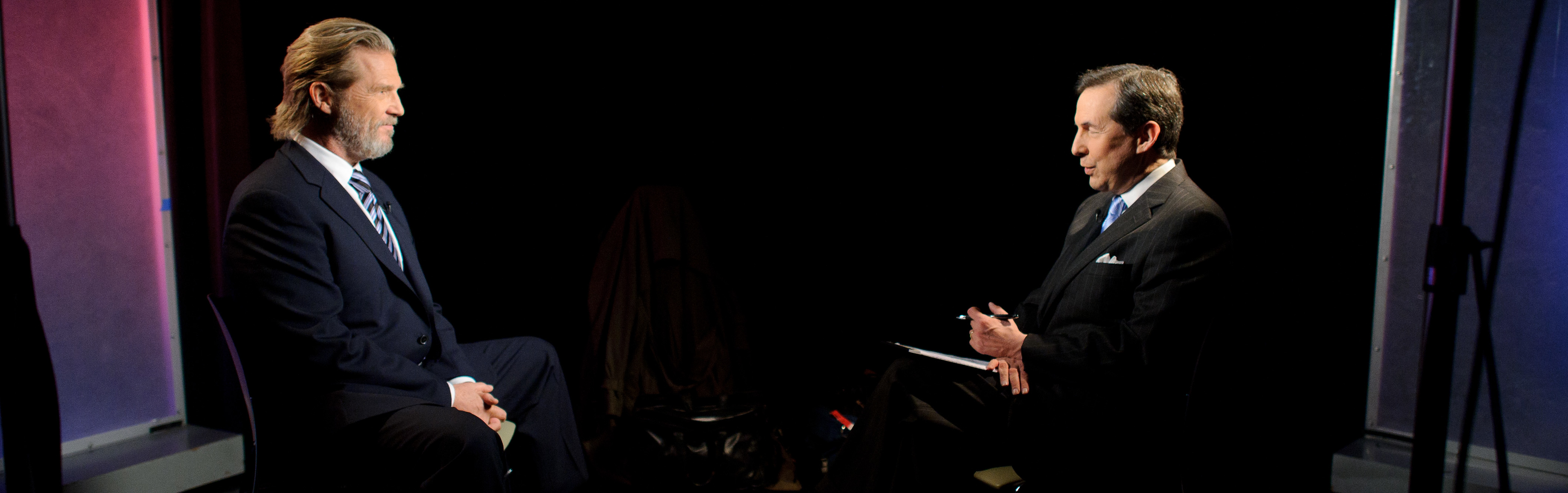 Two men in suits sit facing each other in a dark studio, engaged in a formal interview, with notepads in hand.