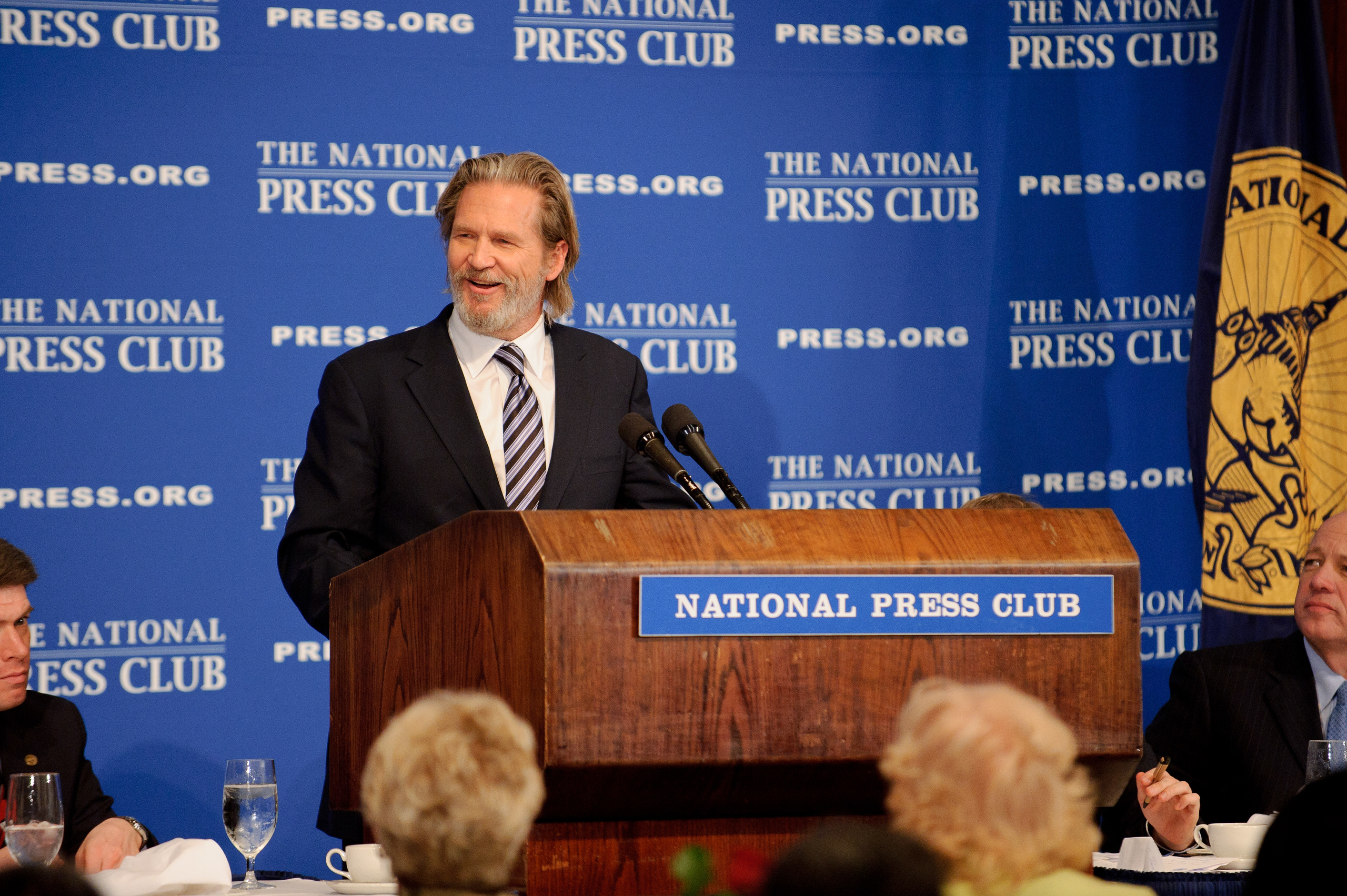 A man in a suit speaks at a podium labeled "National Press Club" with microphones, in front of a blue backdrop and an audience.