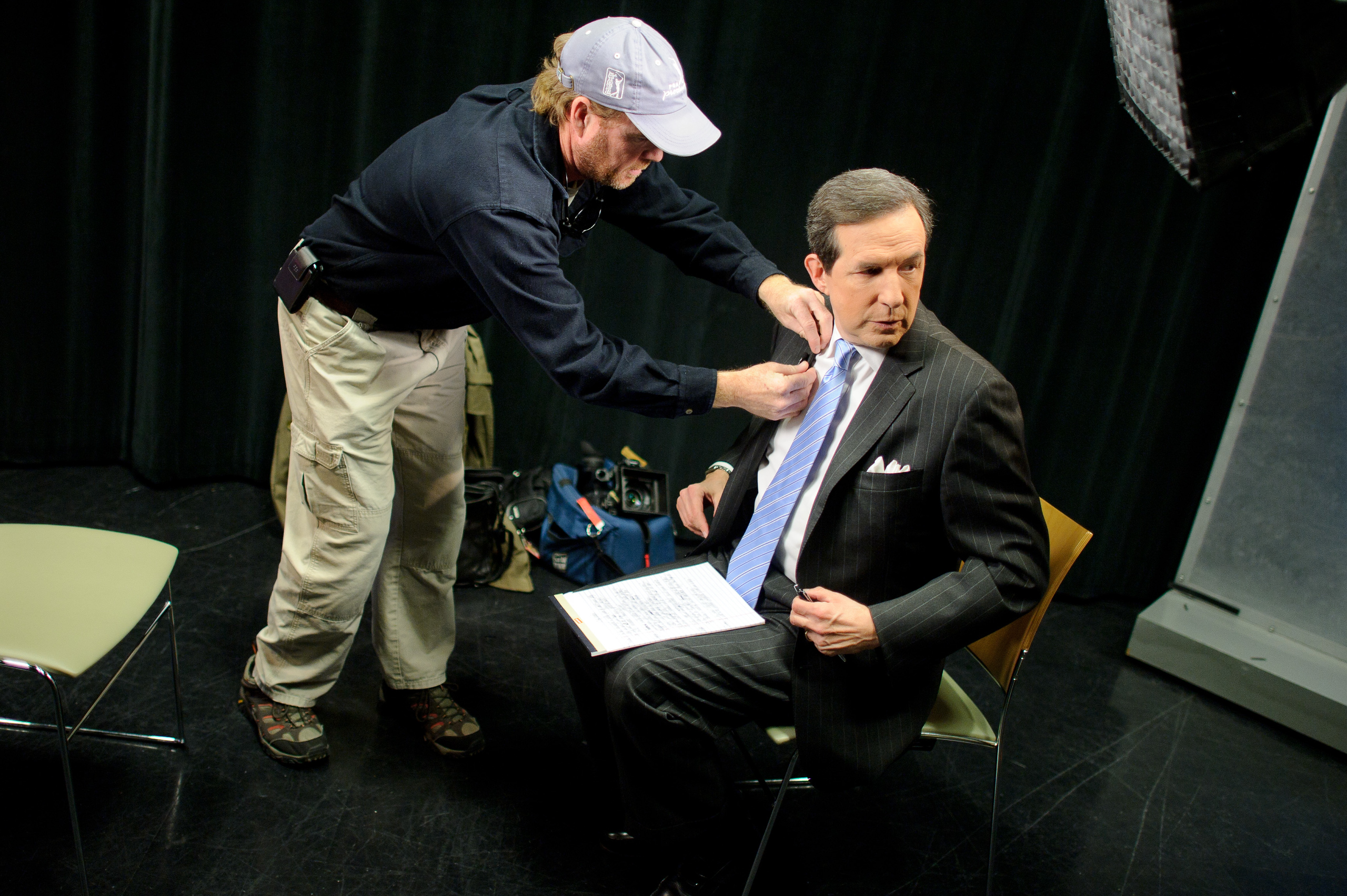 A technician adjusts the microphone on a seated man in a suit holding papers, preparing him for a television or video recording in a studio setting.