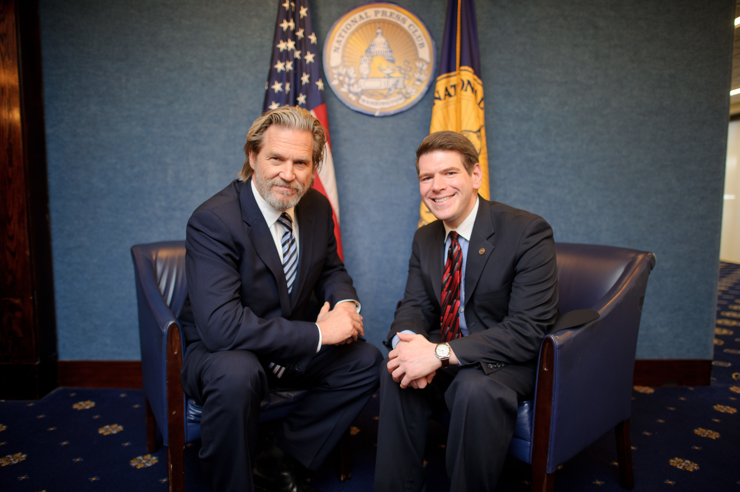 Two men in suits sit in armchairs, posing for a photo in front of American flags and a blue backdrop with official seals.