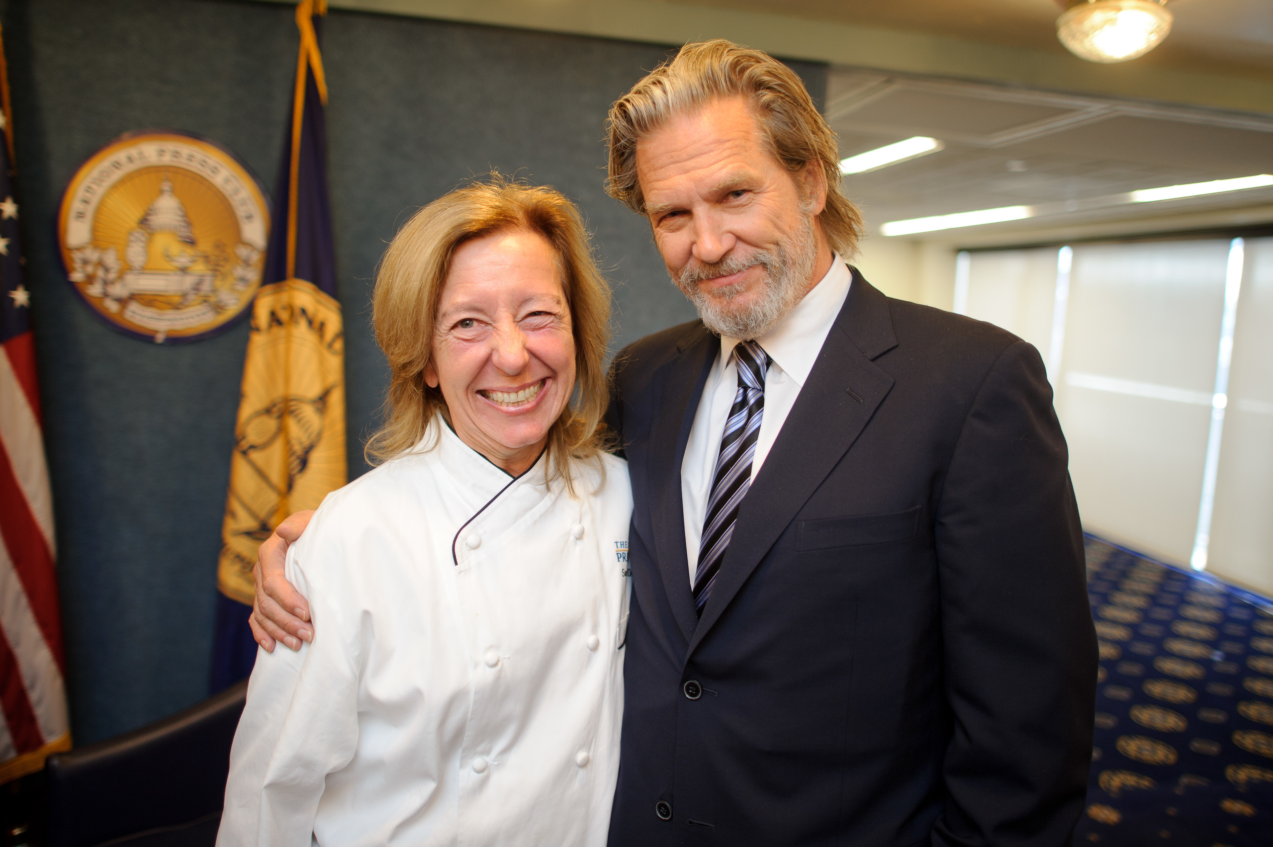 A woman in a chef's coat stands smiling next to a man in a suit and tie, both posing for a photo in a formal room with a flag and emblem in the background.