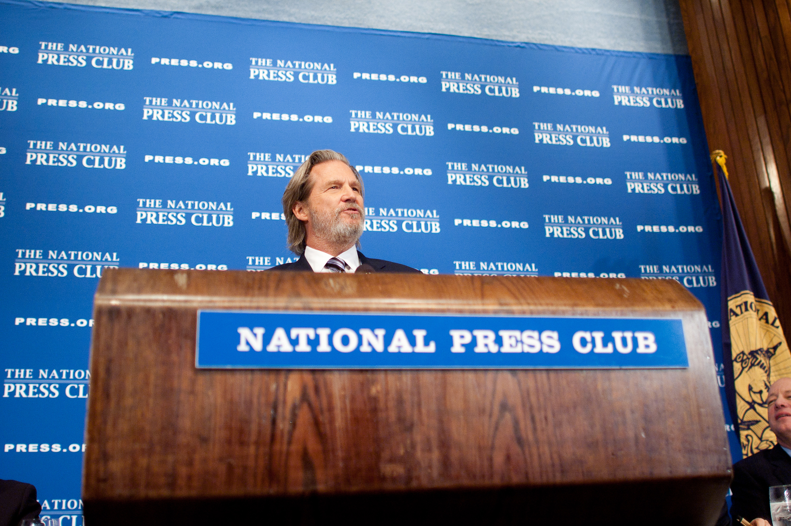 A man in a suit stands at a podium labeled "National Press Club," speaking in front of a blue backdrop with repeated National Press Club logos.