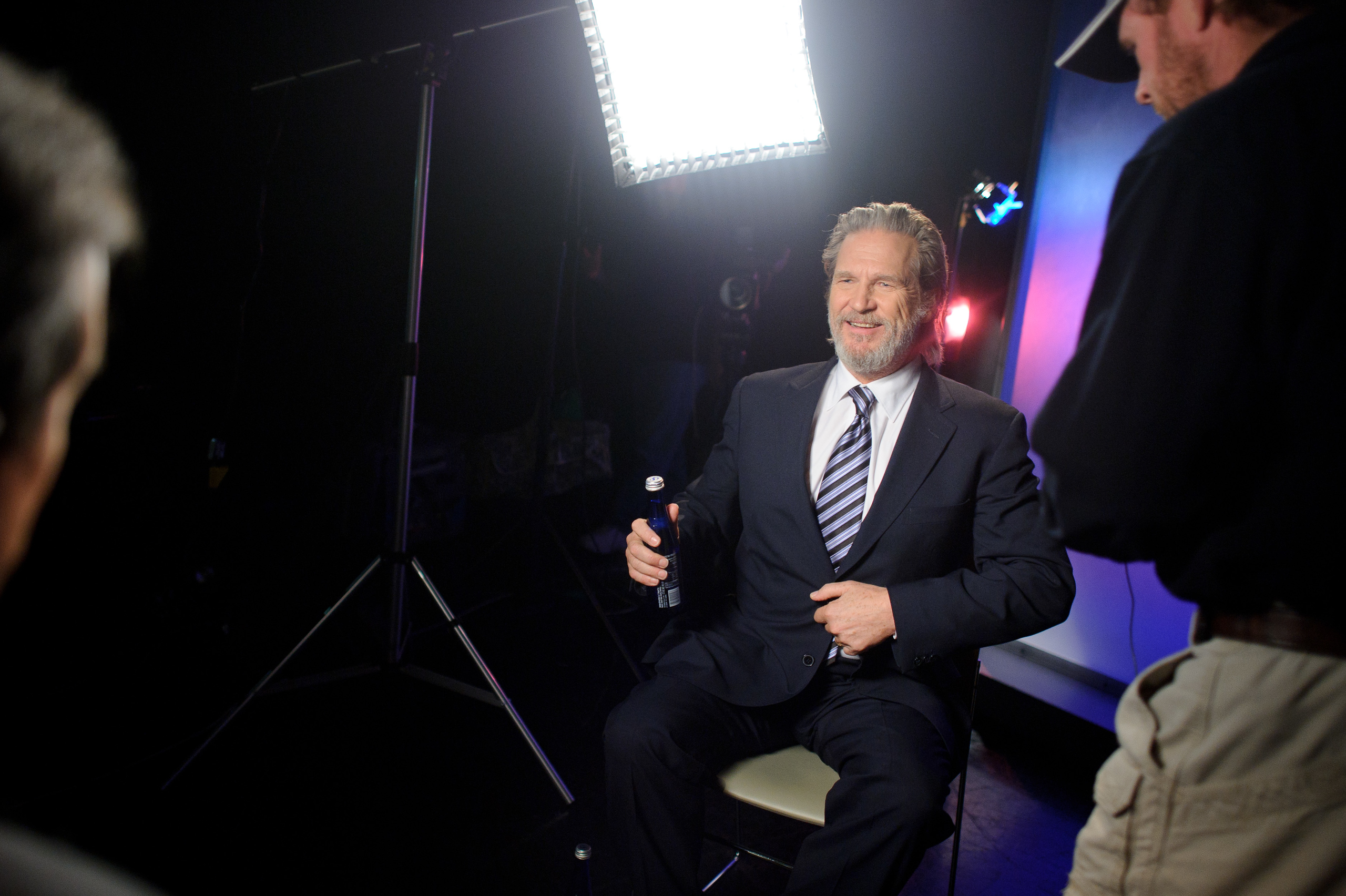 A man in a suit sits on a chair holding a bottle, smiling during a photoshoot with bright studio lights and two other people nearby.