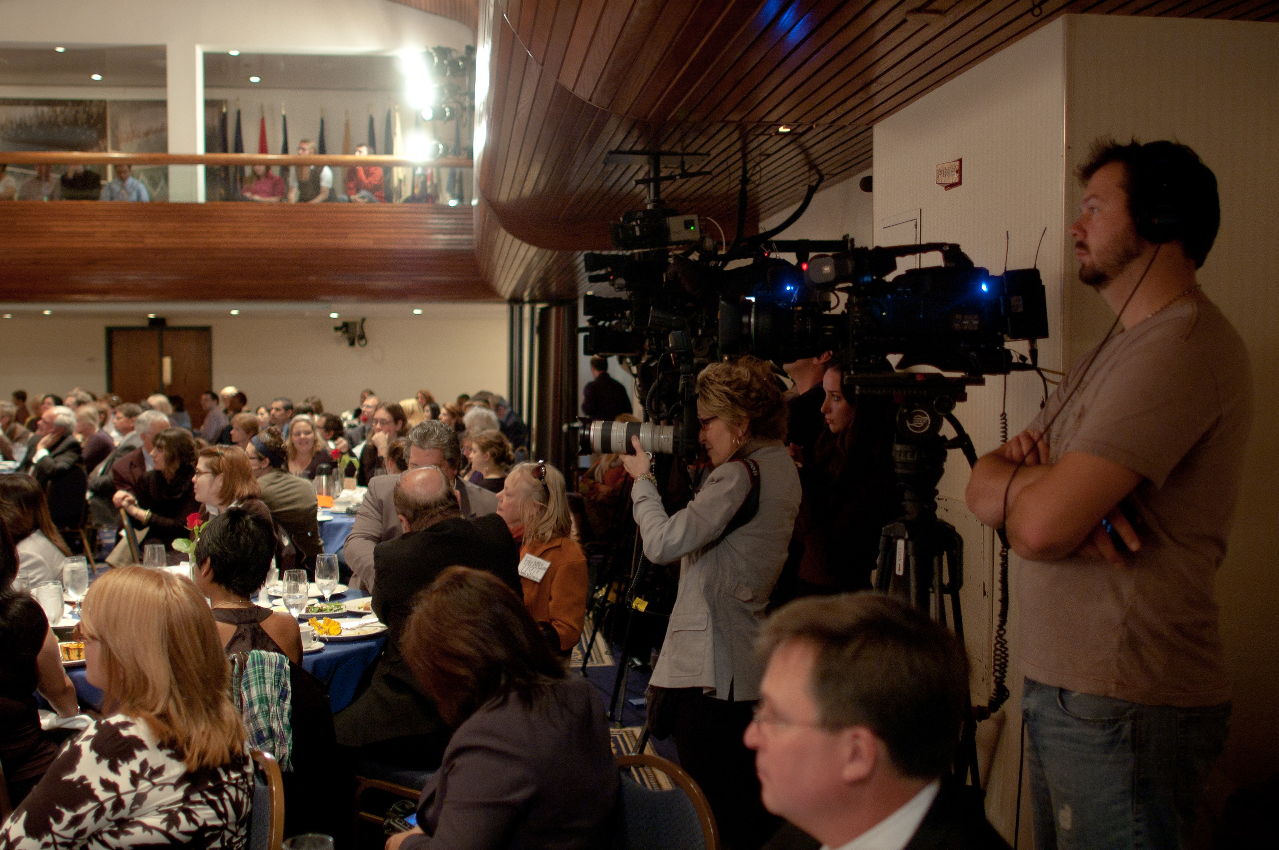 A group of people attend an indoor event while several camera operators film from the side of the room.