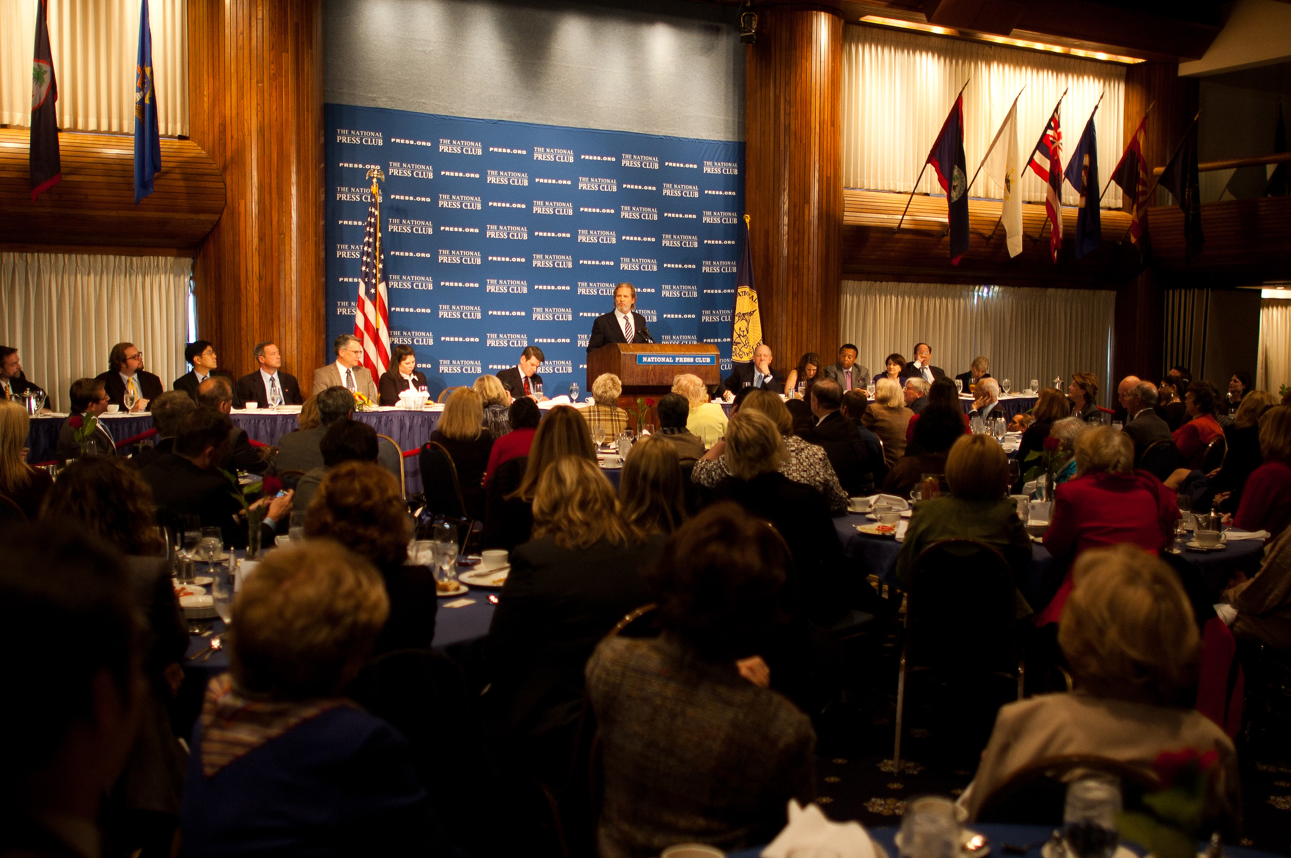 A speaker addresses an audience at a formal event, with people seated at round tables and flags displayed on the walls.
