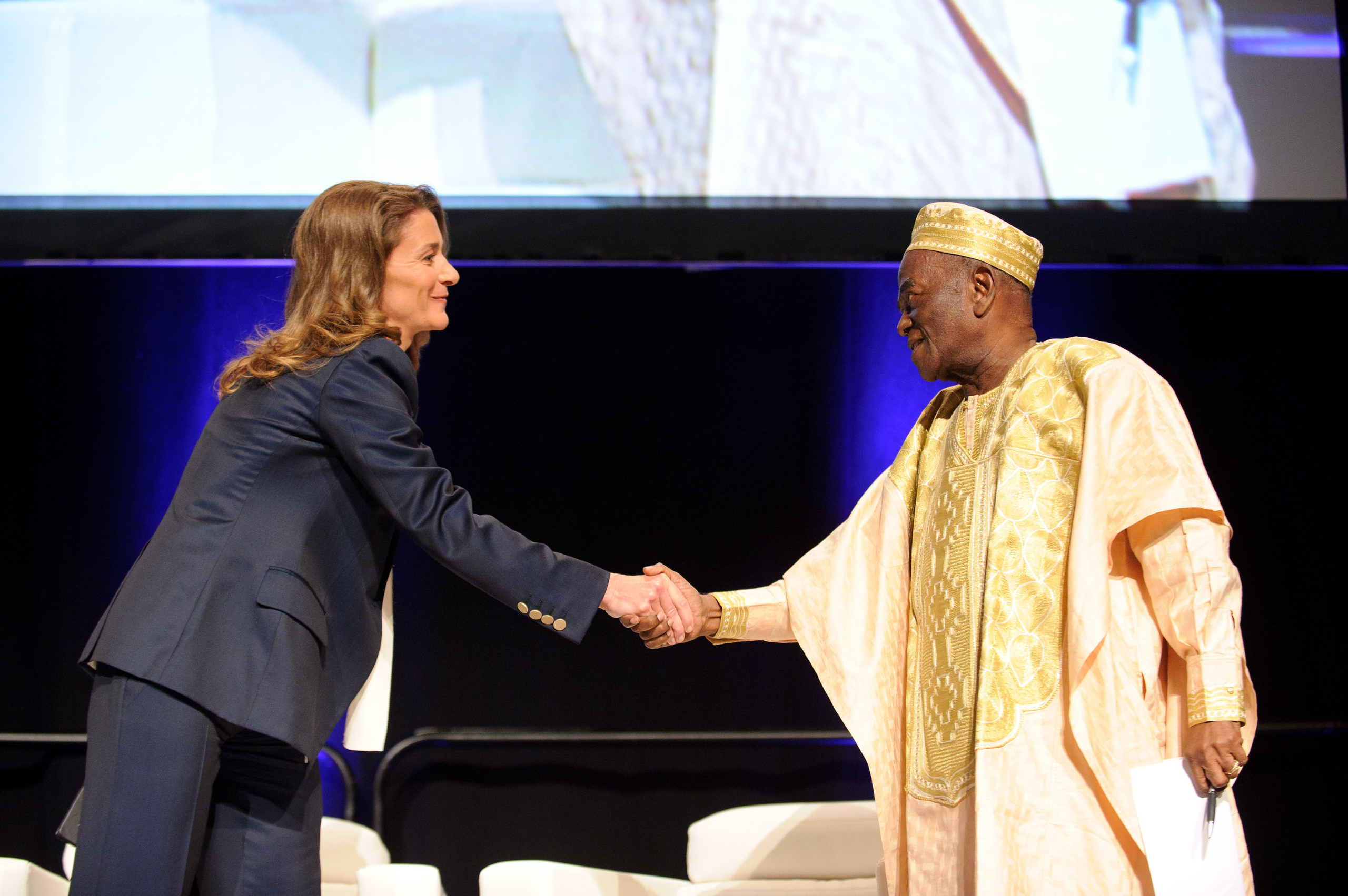 A woman in a business suit and a man in traditional attire shake hands on stage at an event.