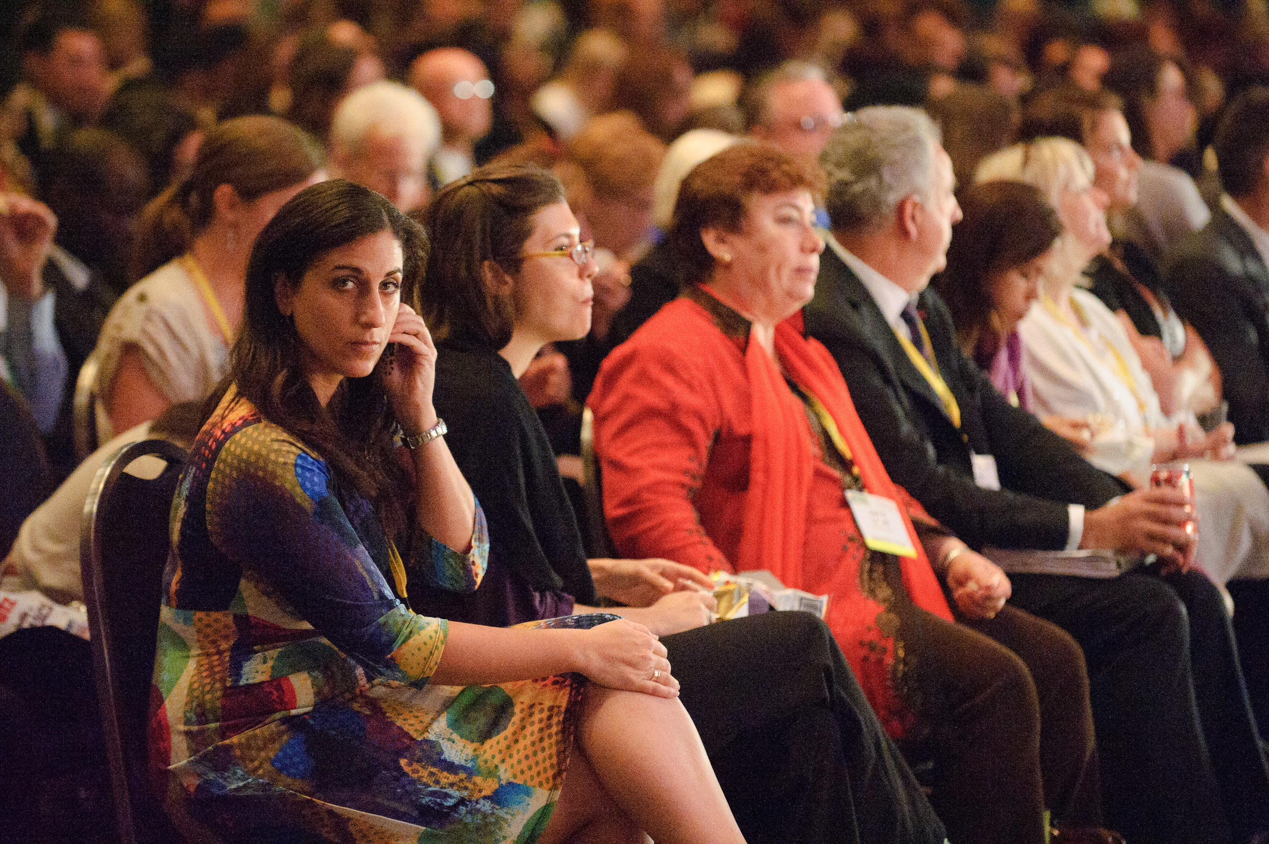 A group of people sit in rows at a conference or event, with one woman in the foreground looking toward the camera.