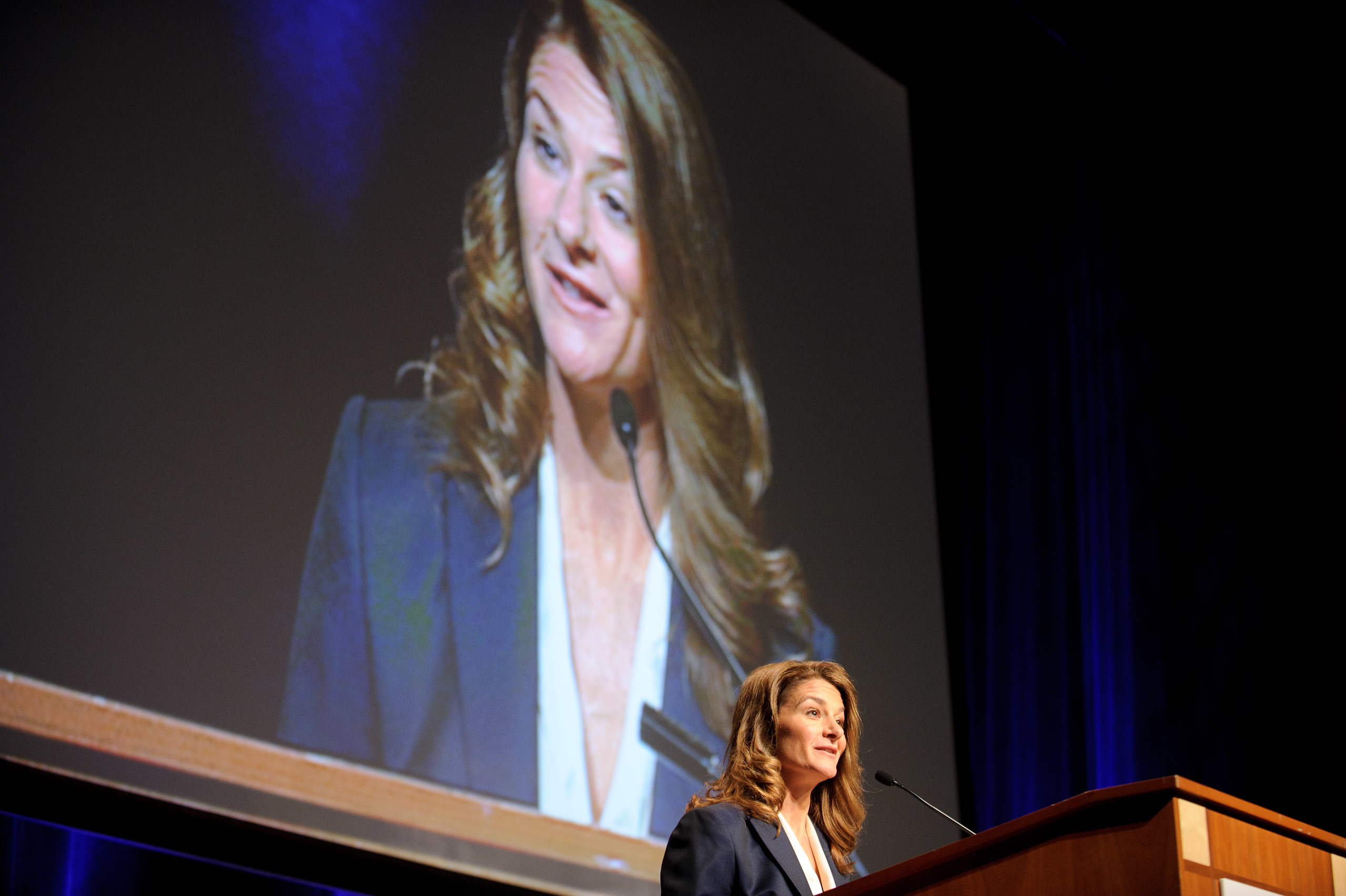 A woman stands at a podium giving a speech, with her image projected on a large screen behind her.