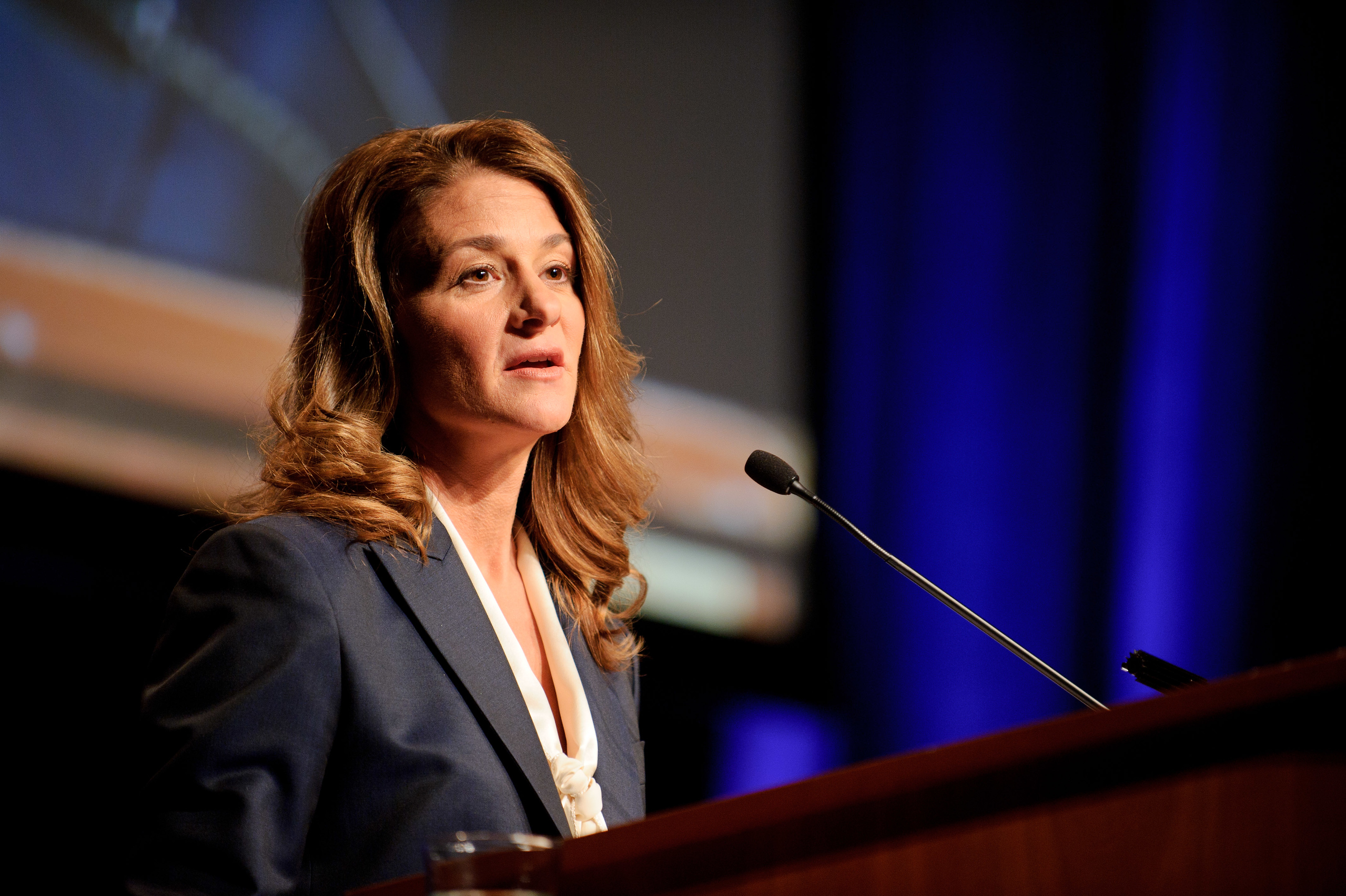 A woman in a suit speaks at a podium with a microphone in front of a dark blue and black background.