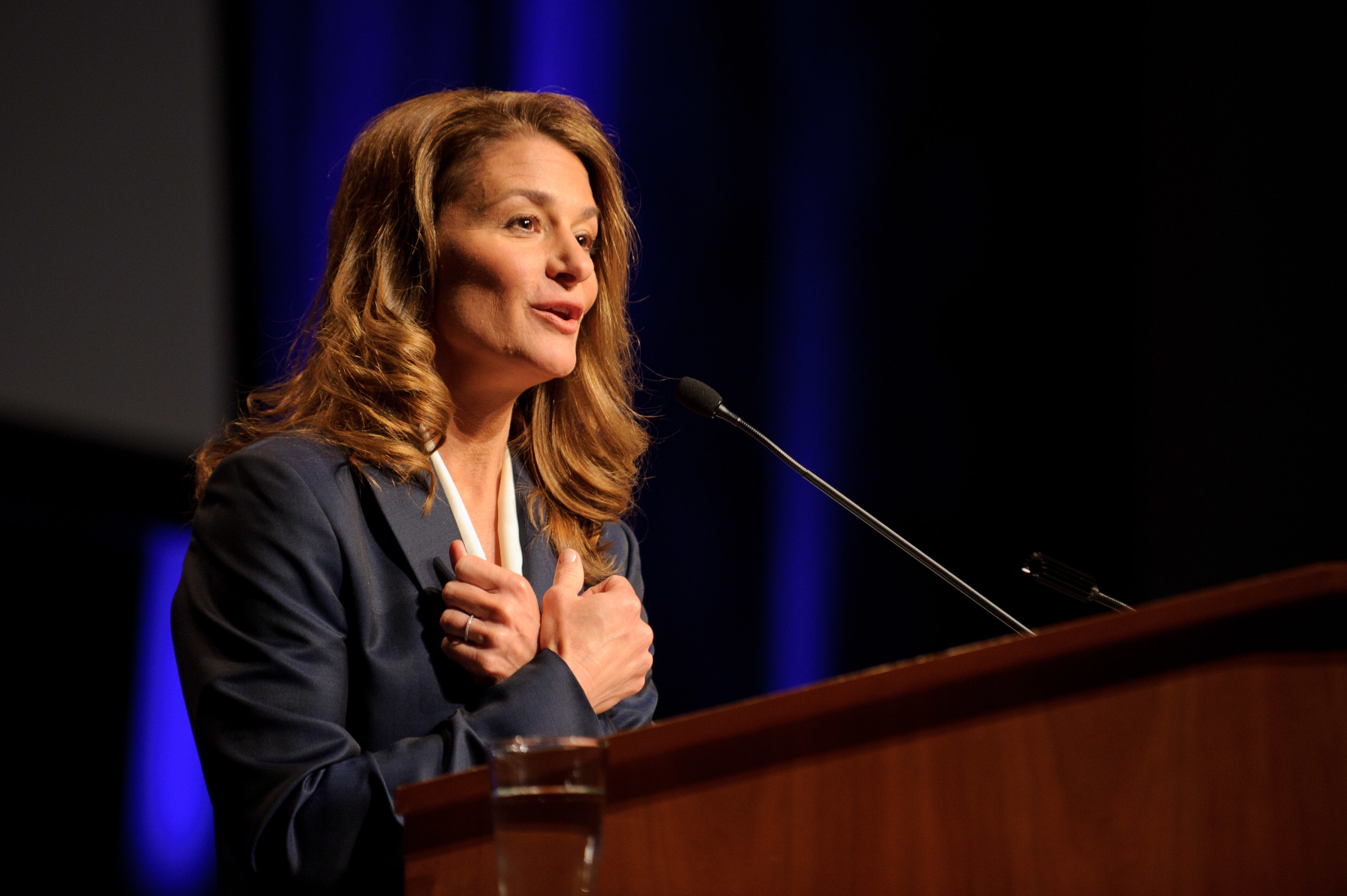 Woman in a suit speaking at a podium with a microphone, blue background, and a glass of water on the podium.