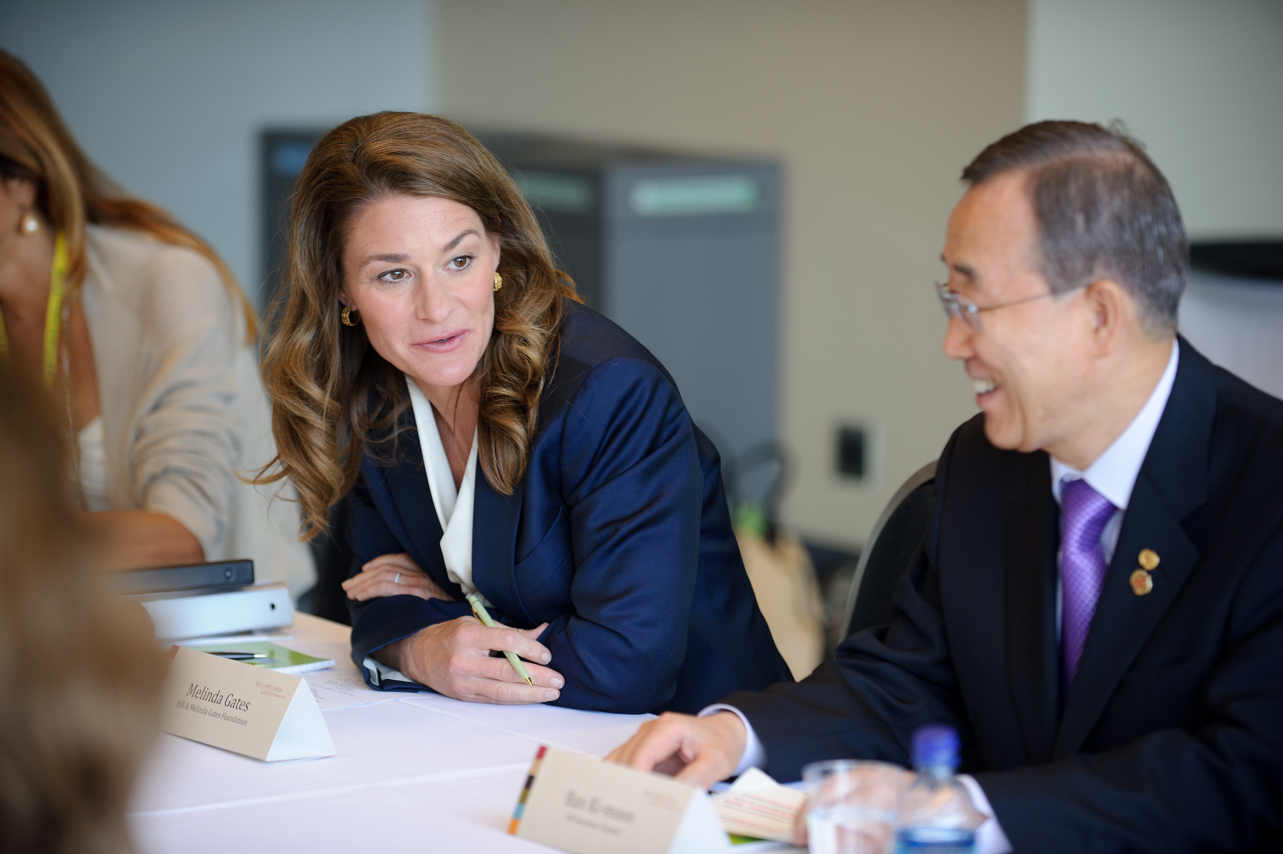 A woman in a navy blazer speaks to a man in a suit and tie at a conference table during a meeting.