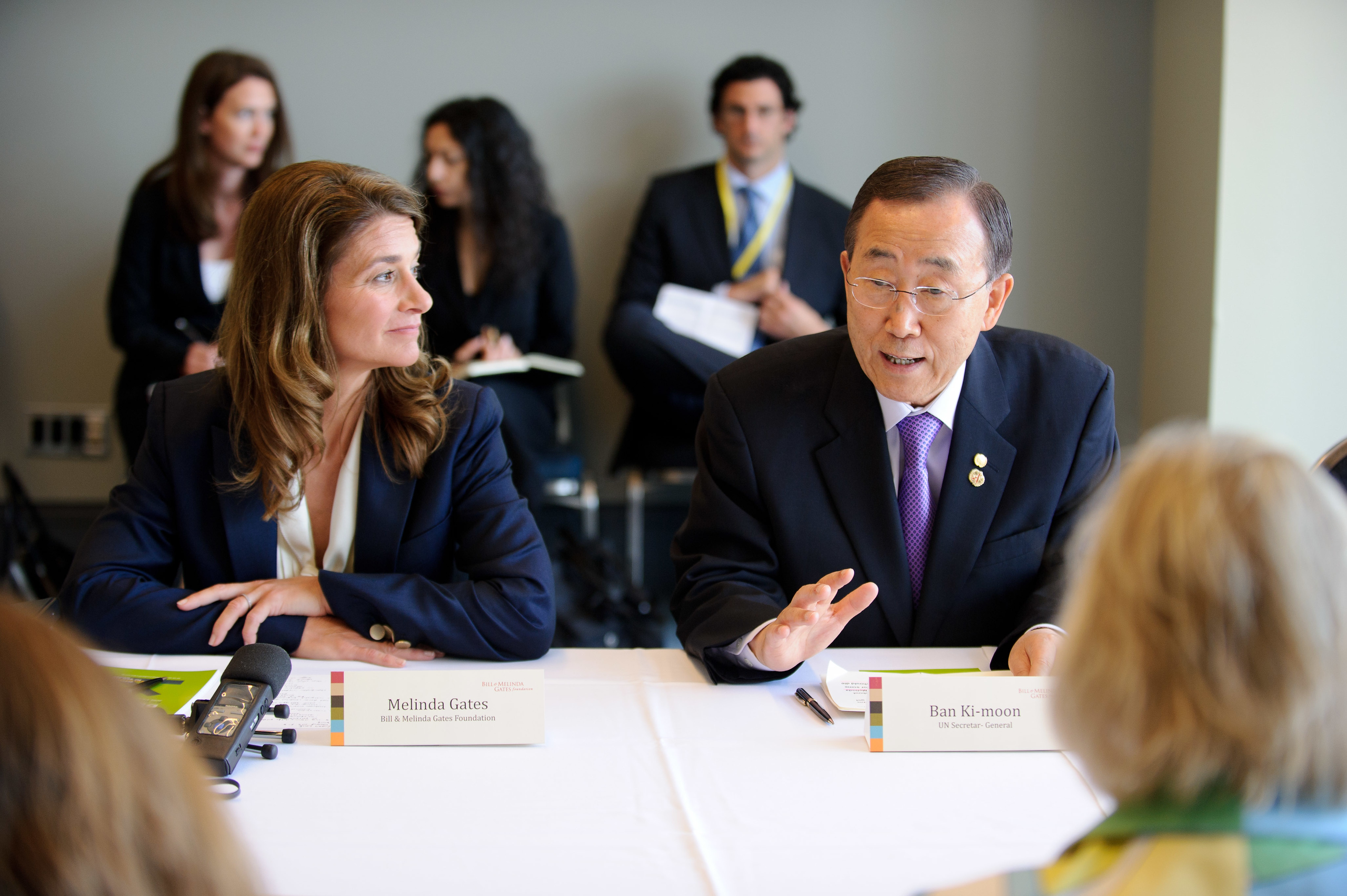 Two people, identified by nameplates as Melinda Gates and Ban Ki-moon, sit at a conference table speaking, with others seated and listening in the background.