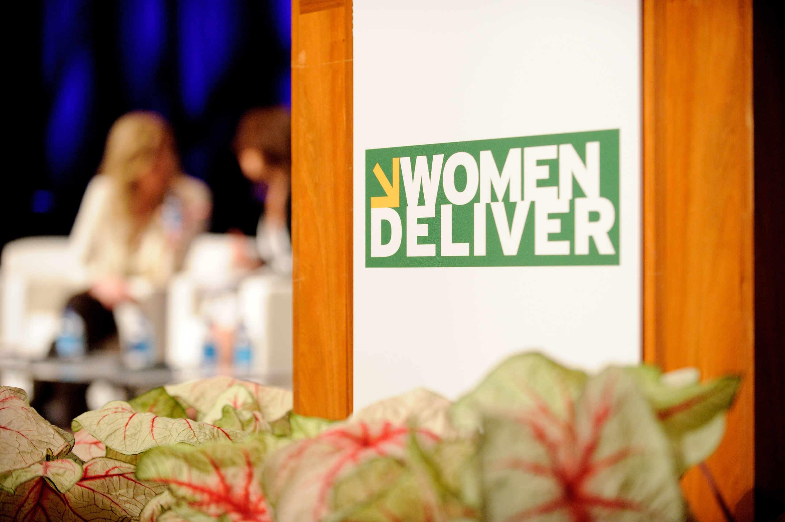 Close-up of a podium with a "Women Deliver" sign, leafy plants in front, and two blurred people sitting in the background.